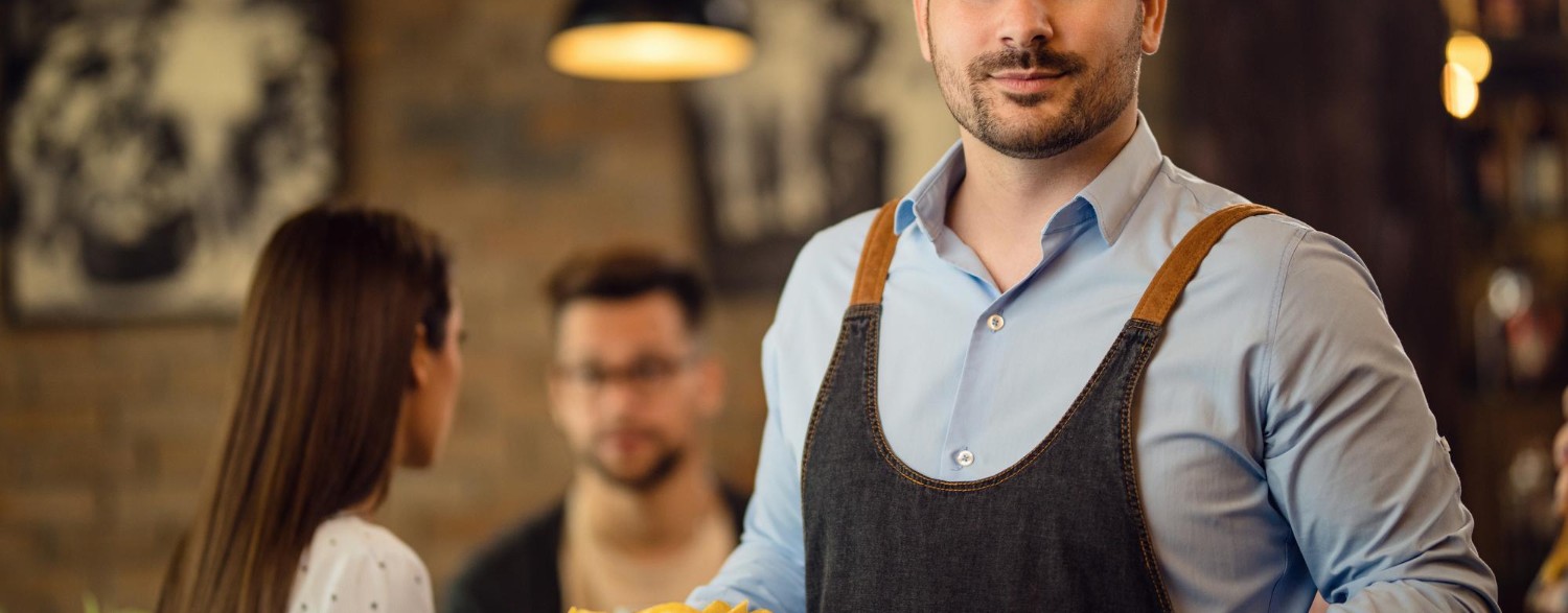 A waiter in a blue shirt and apron holds plates of food, standing in a cozy restaurant with two seated patrons in the background.