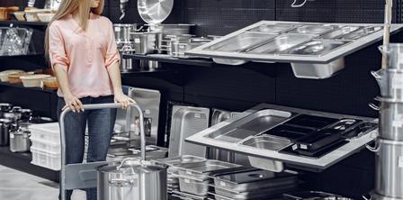 A woman in a pink blouse and jeans pushes a cart with a large pot in a kitchenware store, surrounded by various metal kitchen items.