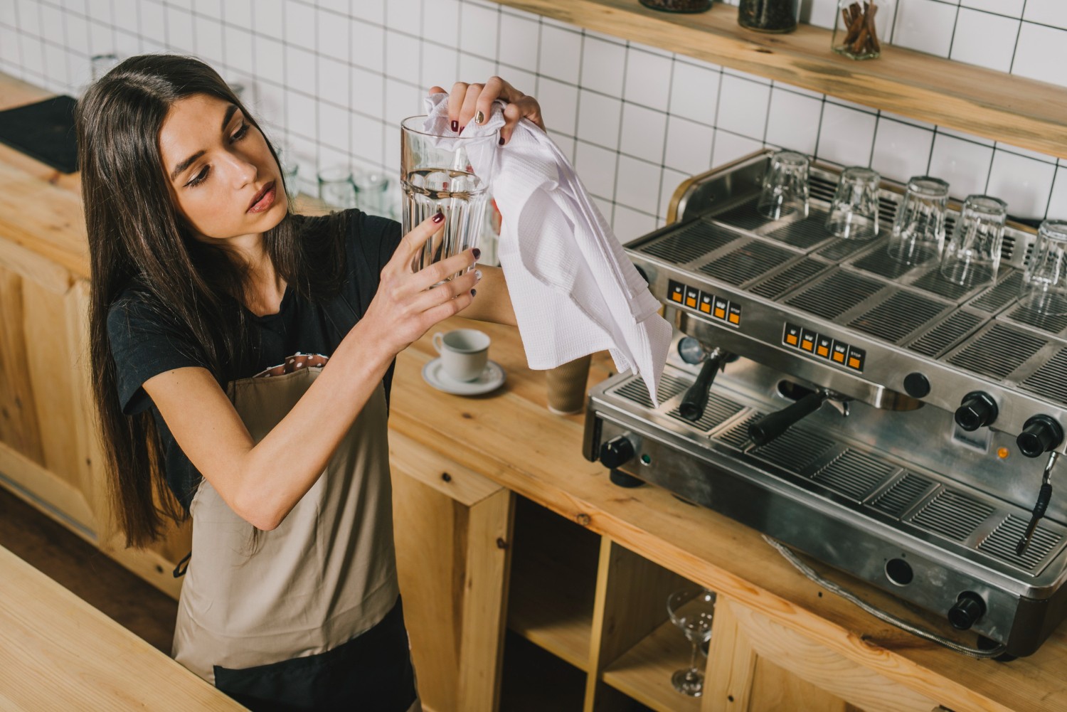 Barista in an apron carefully polishing a glass with a cloth, standing next to a coffee machine in a wooden counter cafe setting.