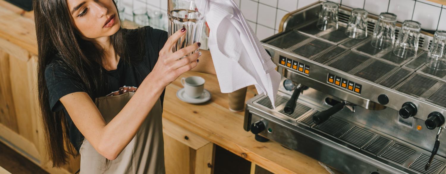Barista in an apron carefully polishing a glass with a cloth, standing next to a coffee machine in a wooden counter cafe setting.