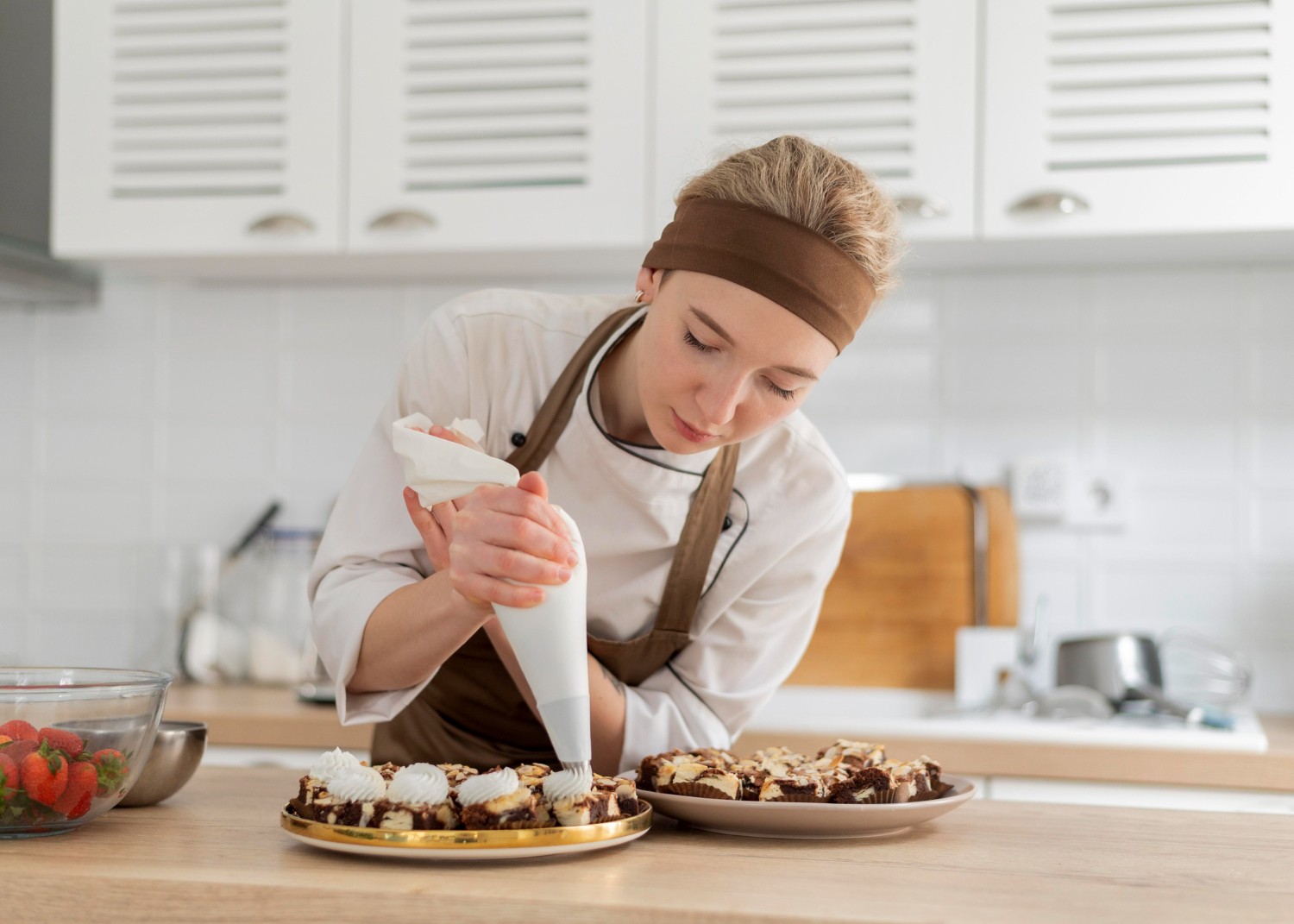 A chef in a white uniform and brown apron decorates pastries with a piping bag in a modern kitchen. A bowl of strawberries is nearby.