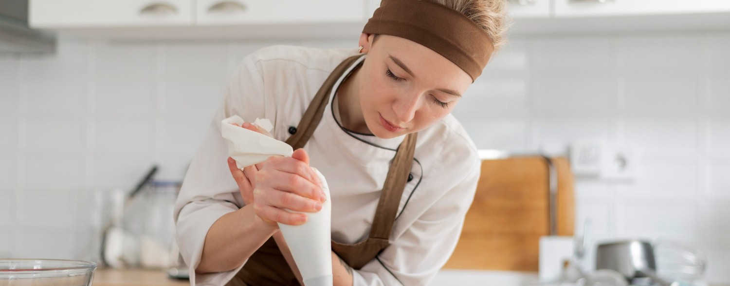 A chef in a white uniform and brown apron decorates pastries with a piping bag in a modern kitchen. A bowl of strawberries is nearby.