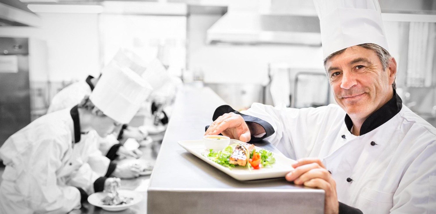 Chef presenting a plated dish with salad and sauce, smiling in a bustling kitchen with other chefs working in the background.