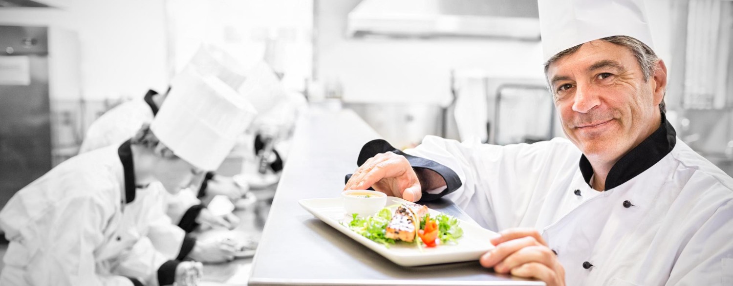 Chef presenting a plated dish with salad and sauce, smiling in a bustling kitchen with other chefs working in the background.