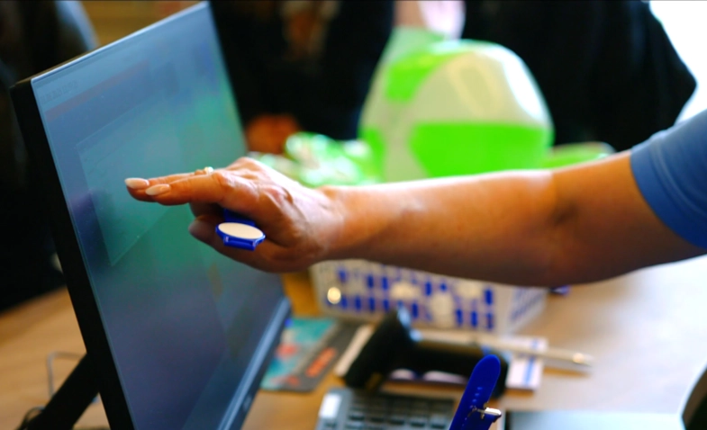 Person, die ein Touchscreen-Display mit einem Finger bedient, umgeben von technischem Zubehör auf einem Schreibtisch.