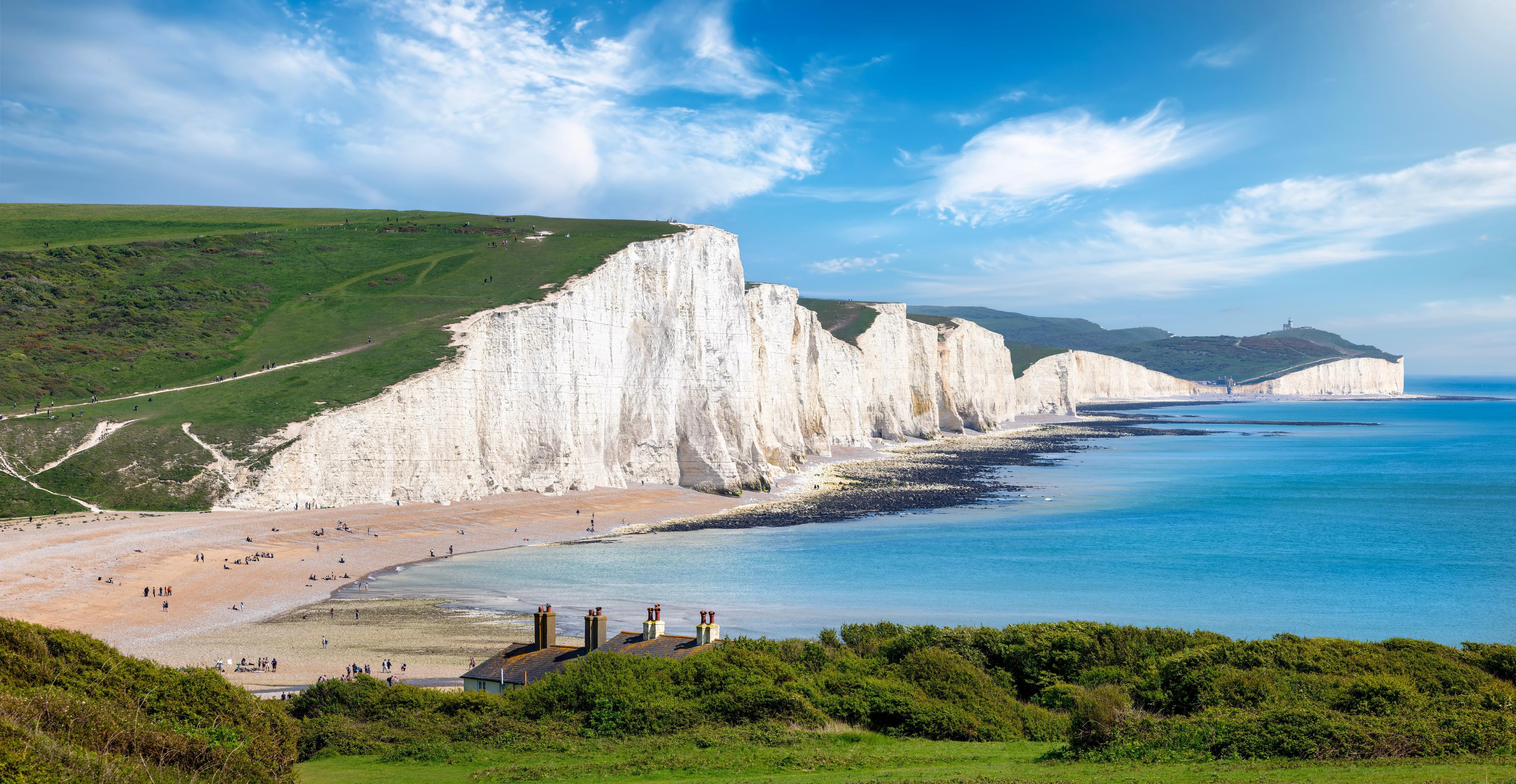 White chalk cliffs of Seven Sisters in England, with green hills above and a pebble beach below, bordering a calm blue sea under a clear sky.