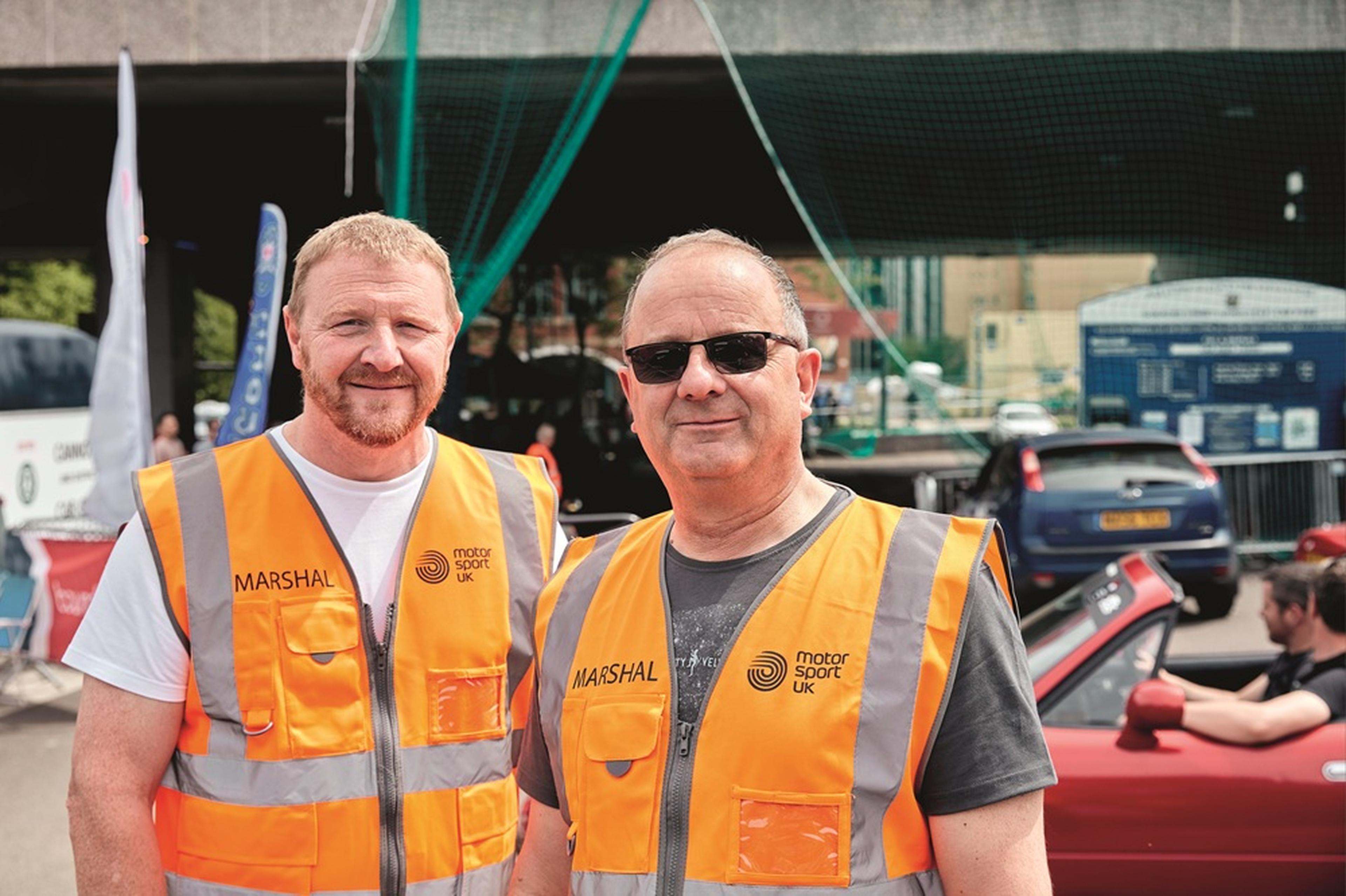 Two men wearing orange safety vests labelled "Marshal" at an outdoor motorsport event