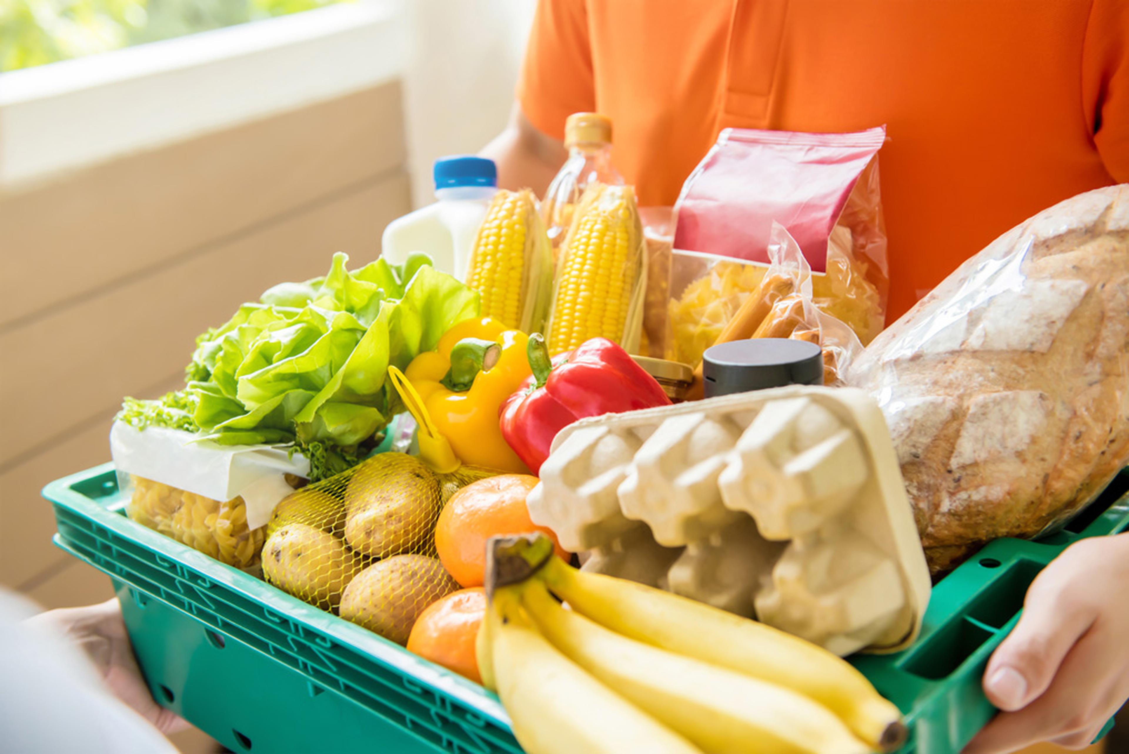 A person holding a crate of fresh groceries, including fruit, vegetables, eggs, bread and milk.