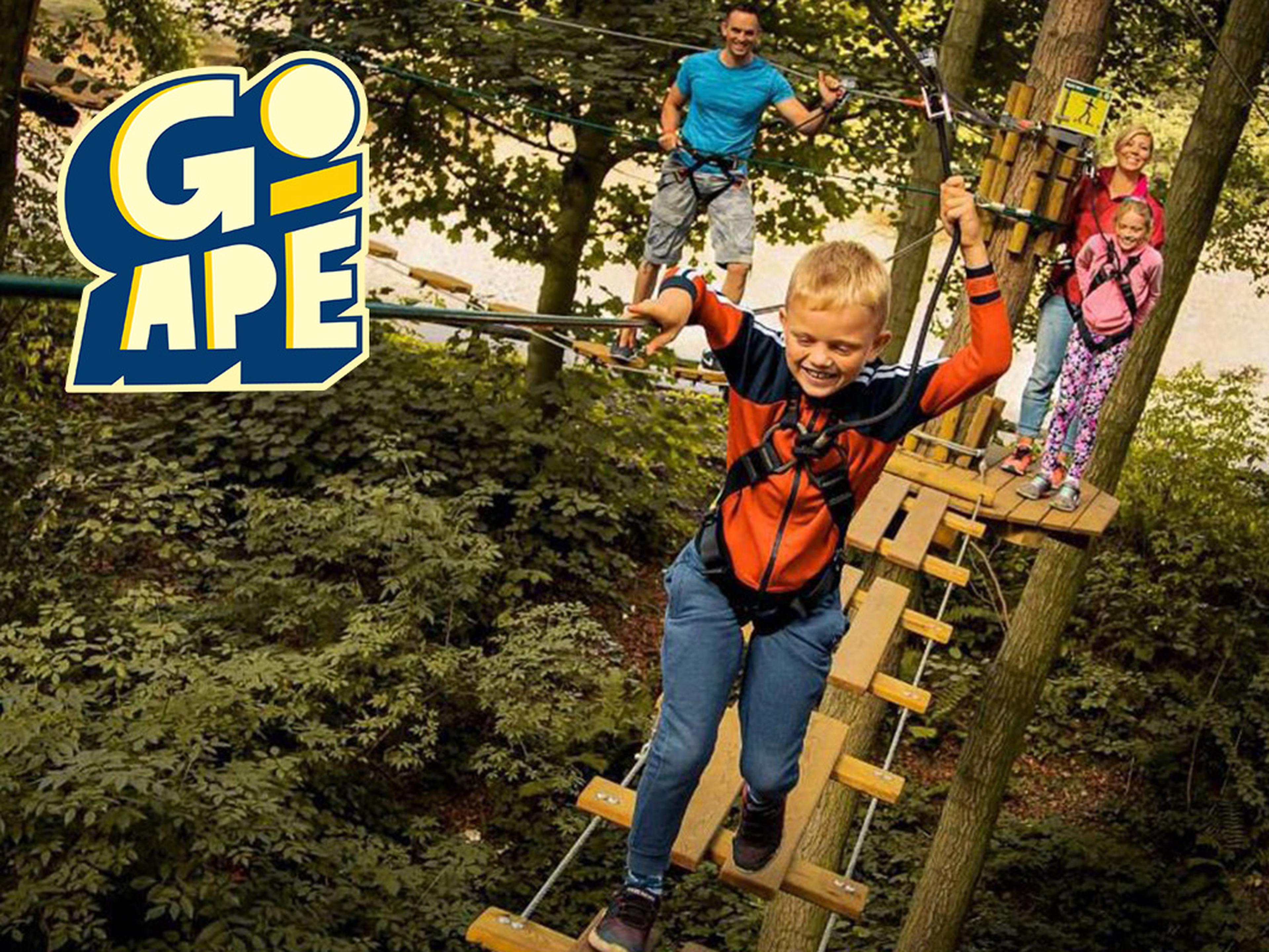 A boy navigates a treetop rope course with family, surrounded by lush greenery. "Go Ape" logo is visible in the corner.