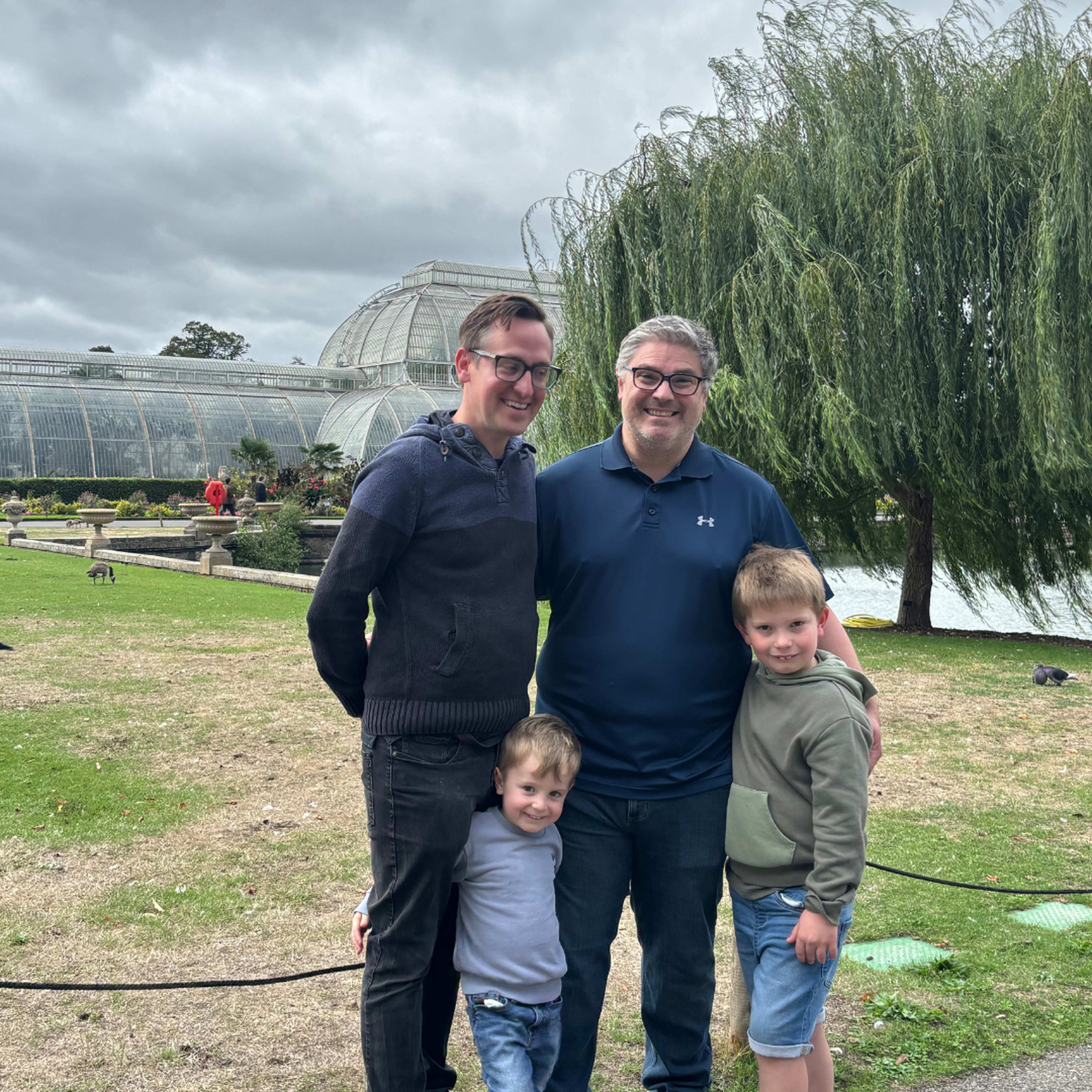 Two smiling men and two young boys stand together on a lawn at Kew Gardens. Behind them is the glass Palm House, with ornamental stone urns along a path, and on the right a weeping willow hangs over a lake under a cloudy grey sky.