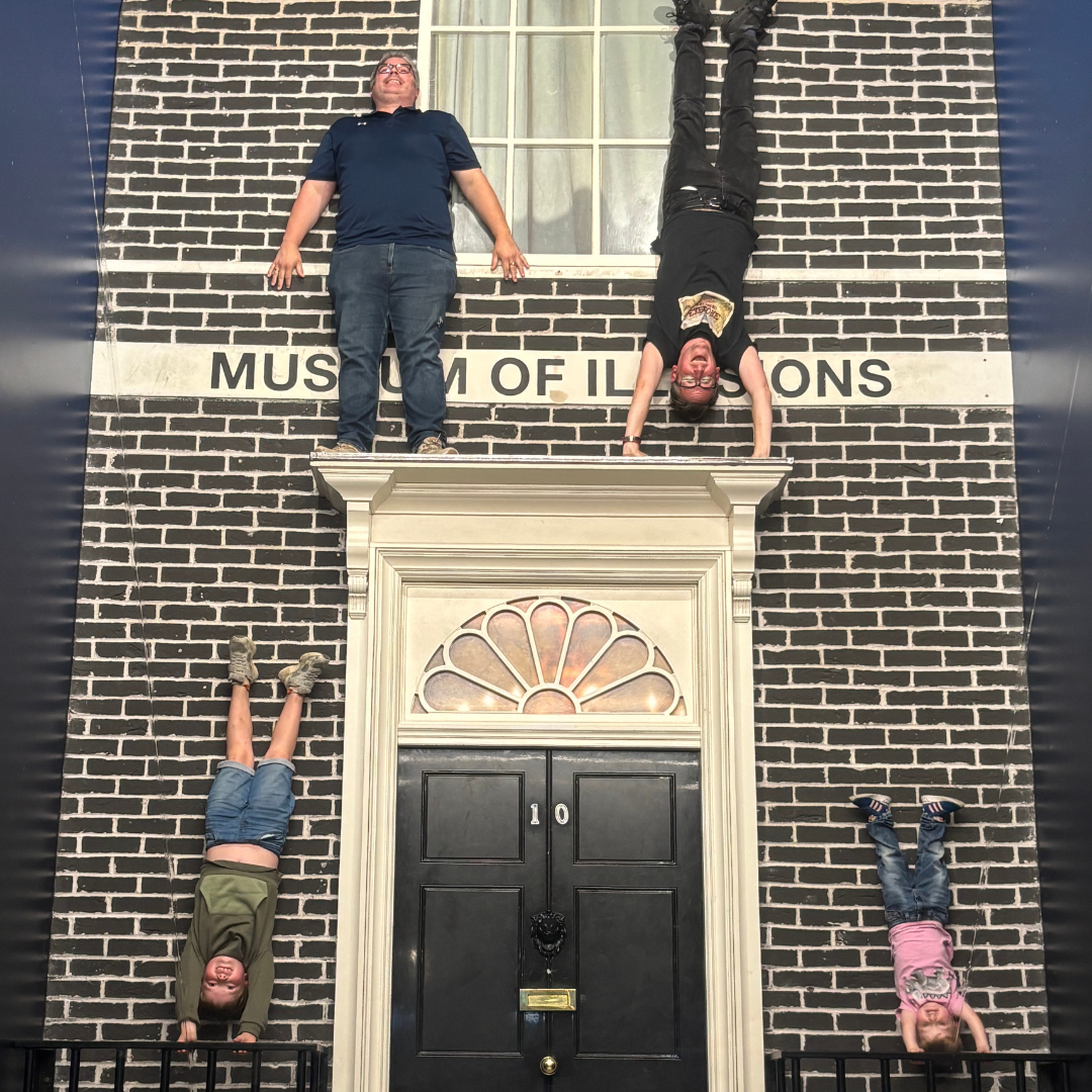 An optical illusion room at the Museum of Illusions London, with dark blue walls covered in bright blue and teal chevrons. On the left, a man appears to tower over a small boy in a pink T-shirt. In the far right corner, another man holding the older boy looks tiny by comparison, though the room's angled shape creates the size difference.