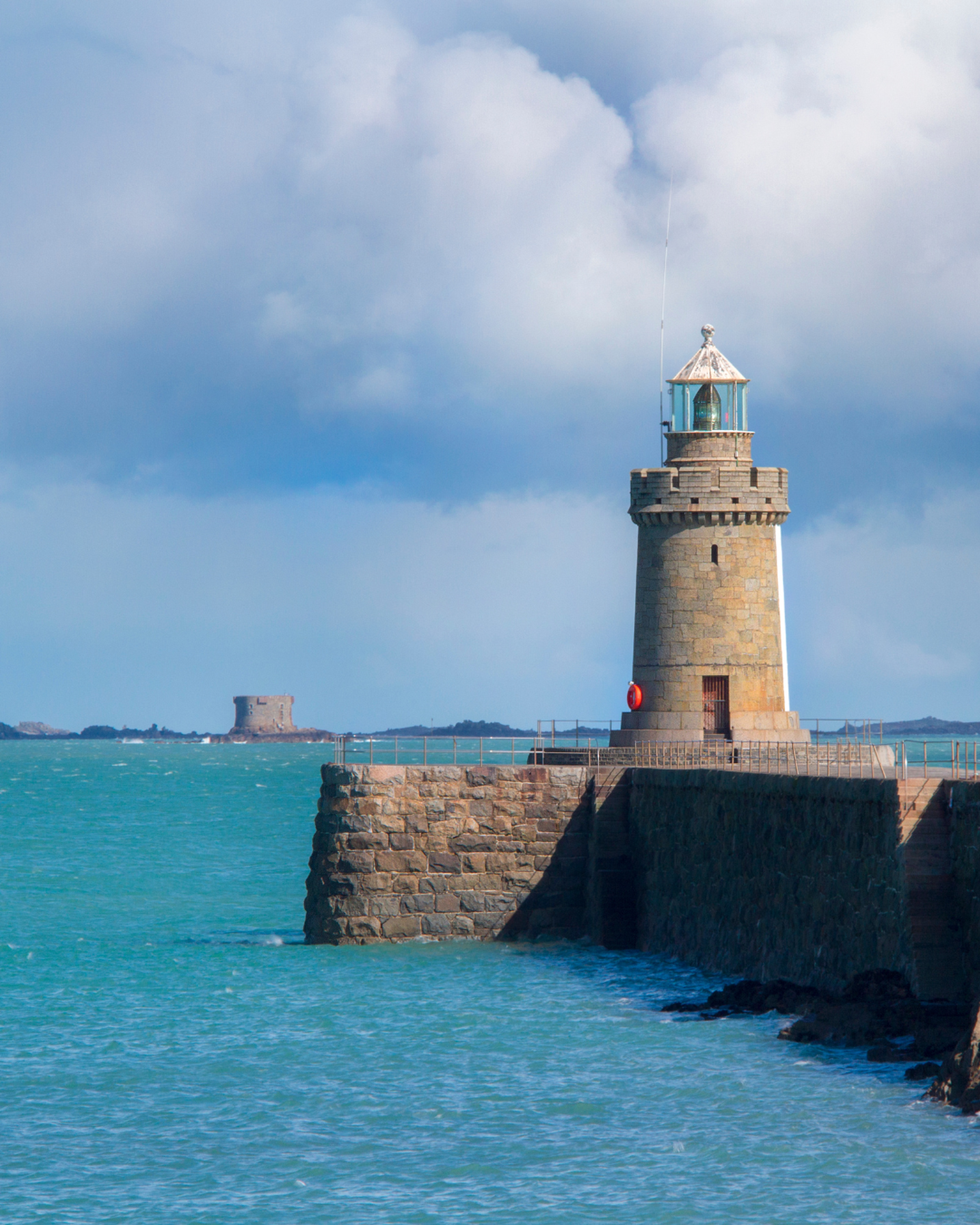 A stone lighthouse stands on a rocky pier by turquoise waters, with a cloudy sky and distant land in the background.