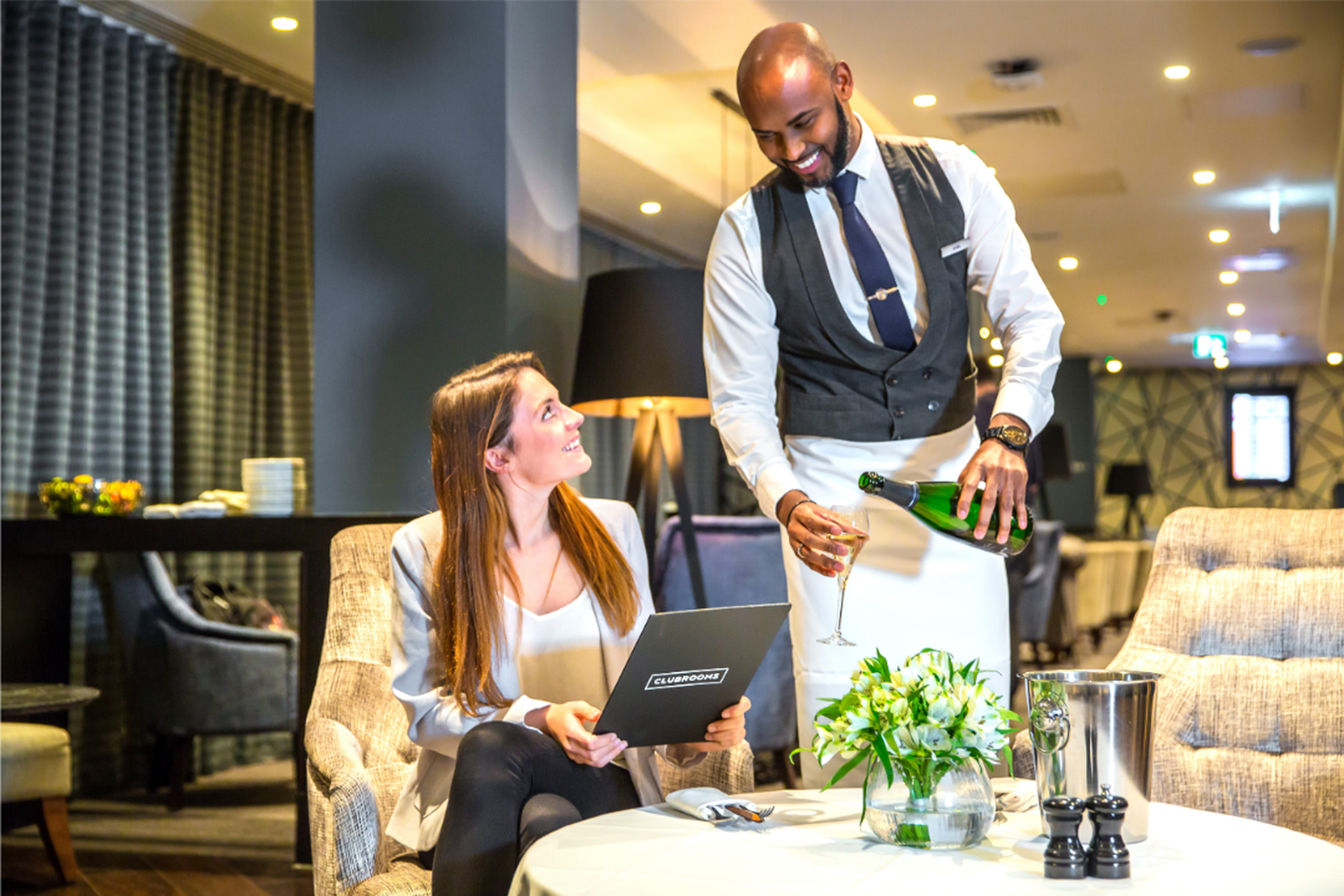 Server pouring champagne for a smiling woman with a laptop in a cozy, elegant airport lounge..