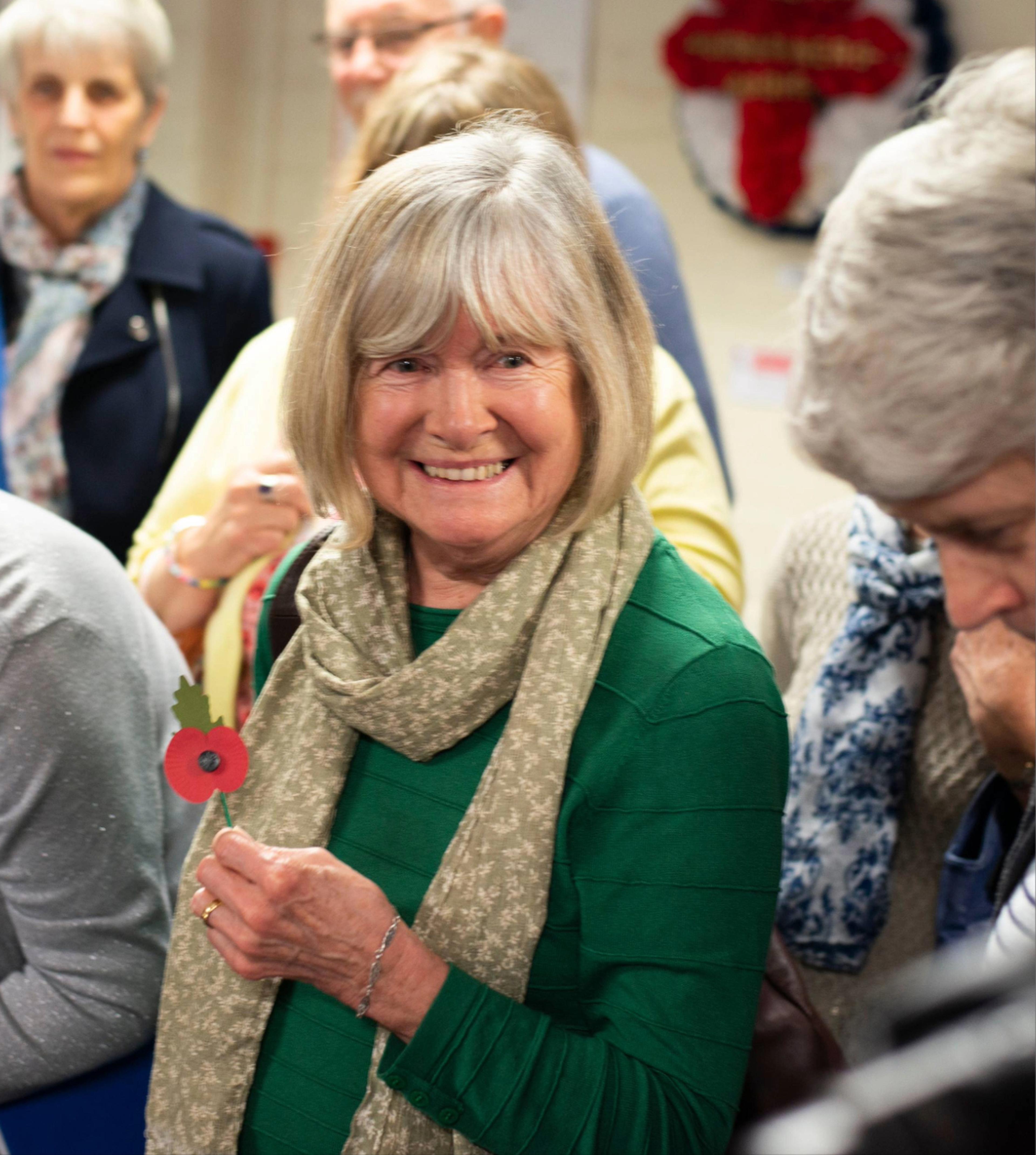 Member in a green sweater and scarf smiles, holding a red poppy paper flower, surrounded by people indoors.
