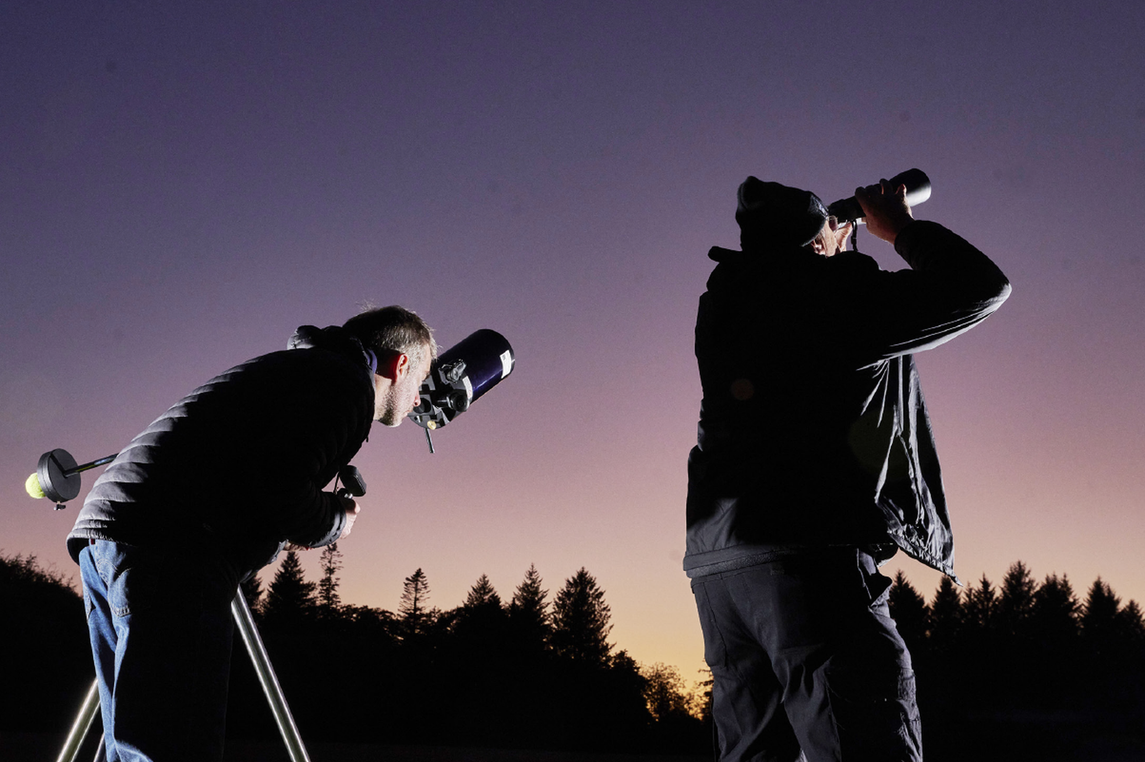Two people stargazing at dusk, one using a telescope and the other binoculars, with trees silhouetted against the sky.