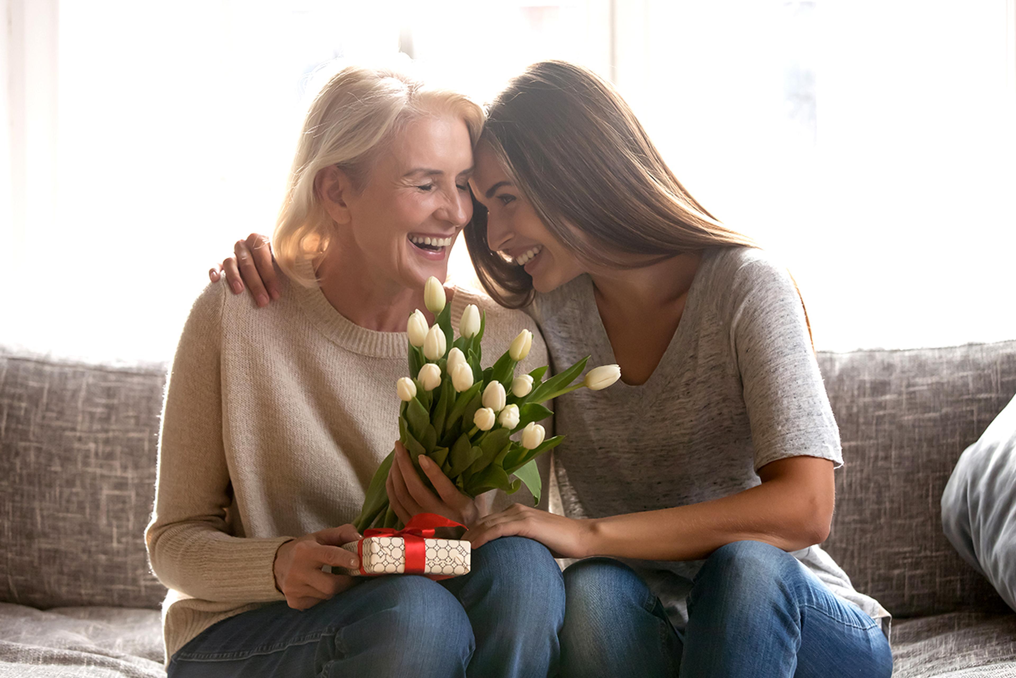 Two women sitting on a couch share a joyful moment. One holds a bouquet of white tulips and a small gift box. They are smiling and leaning close together.