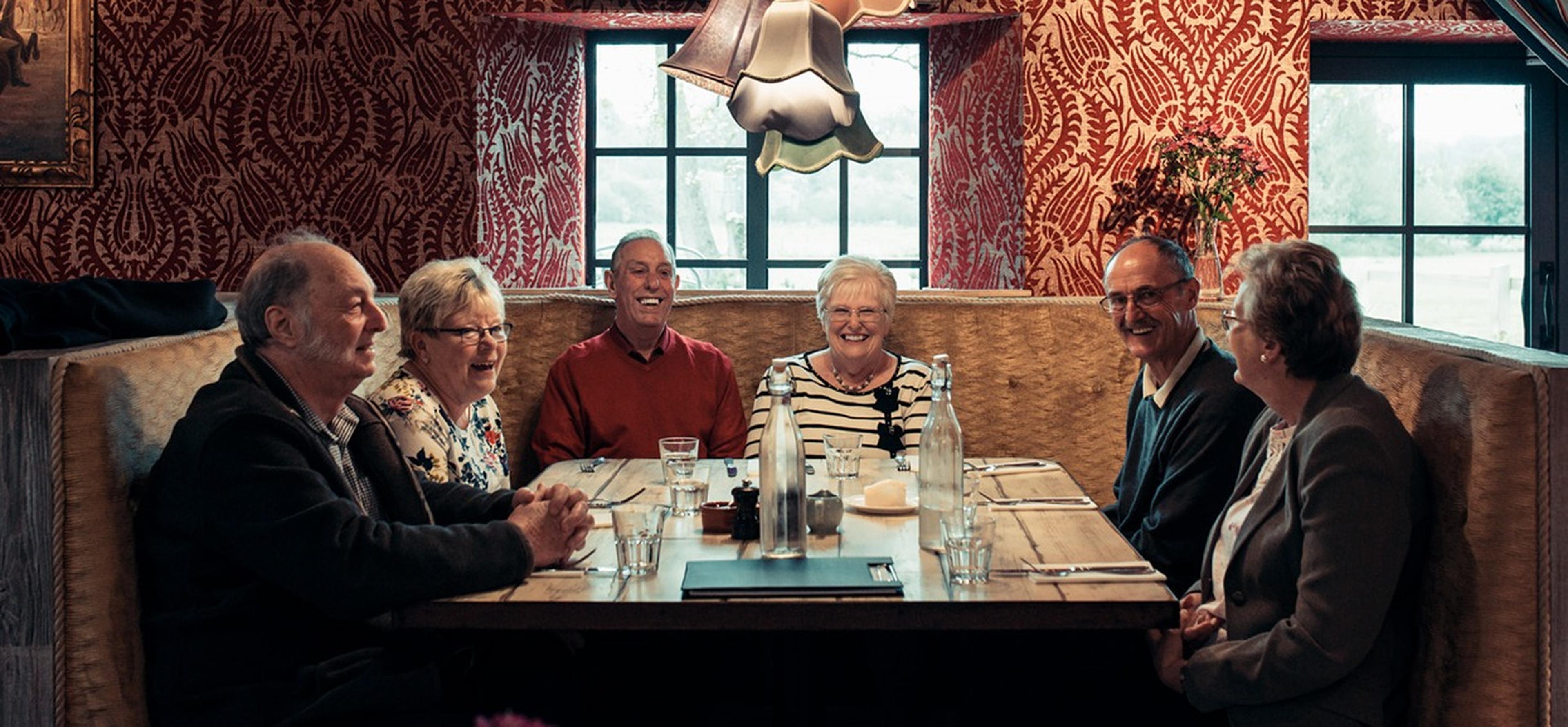 A group of six elderly people sitting around a wooden table in a cozy restaurant with red patterned walls, smiling and conversing.