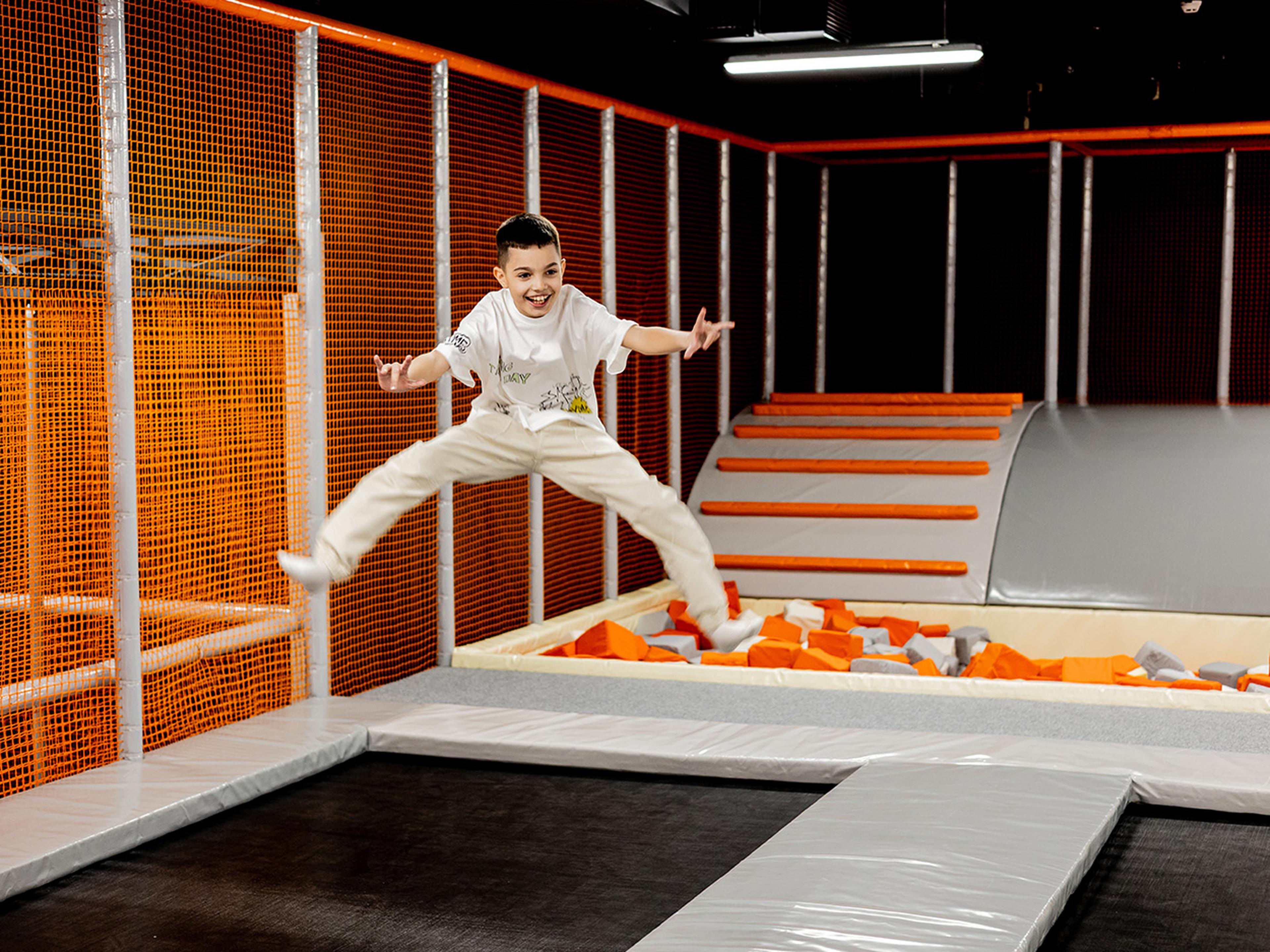 A child jumps energetically on a trampoline, surrounded by orange netting and a foam pit in an indoor play area.
