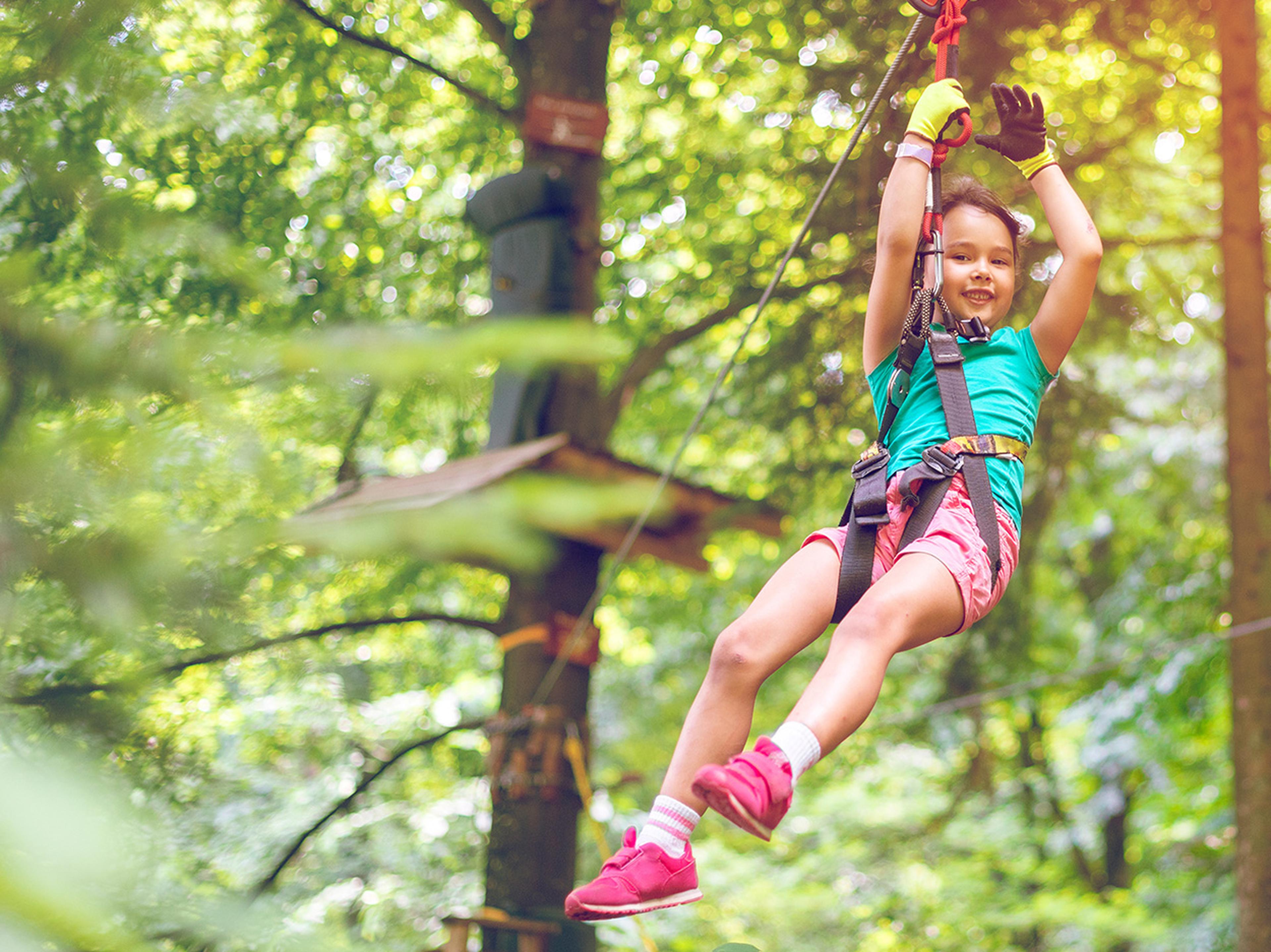 Child in safety gear joyfully ziplining through a lush forest, wearing pink shoes and a blue shirt, with sunlight filtering through the trees.