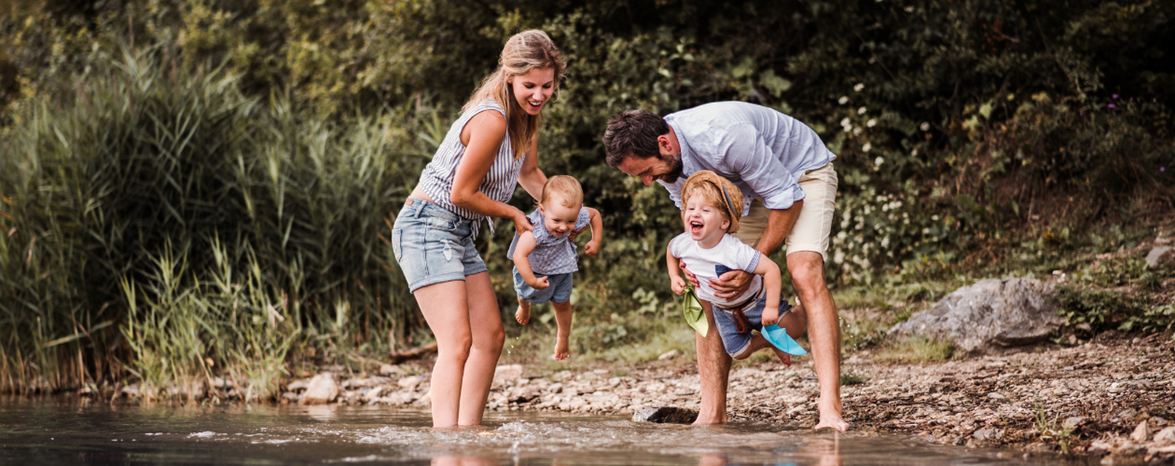A family of four playing by a lakeside, with parents holding their young children above the water on a rocky shoreline.