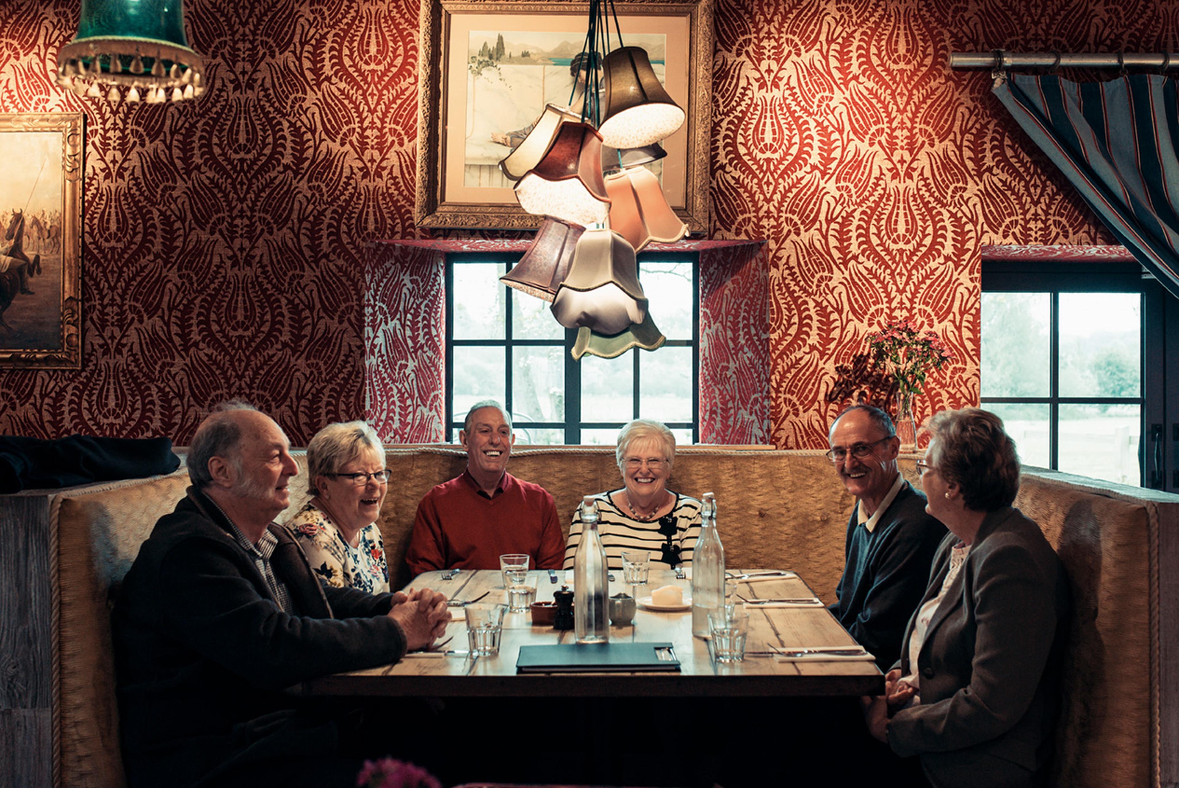 Six people smiling and sitting around a wooden table in a cozy room with red-patterned wallpaper and decorative lamps above.