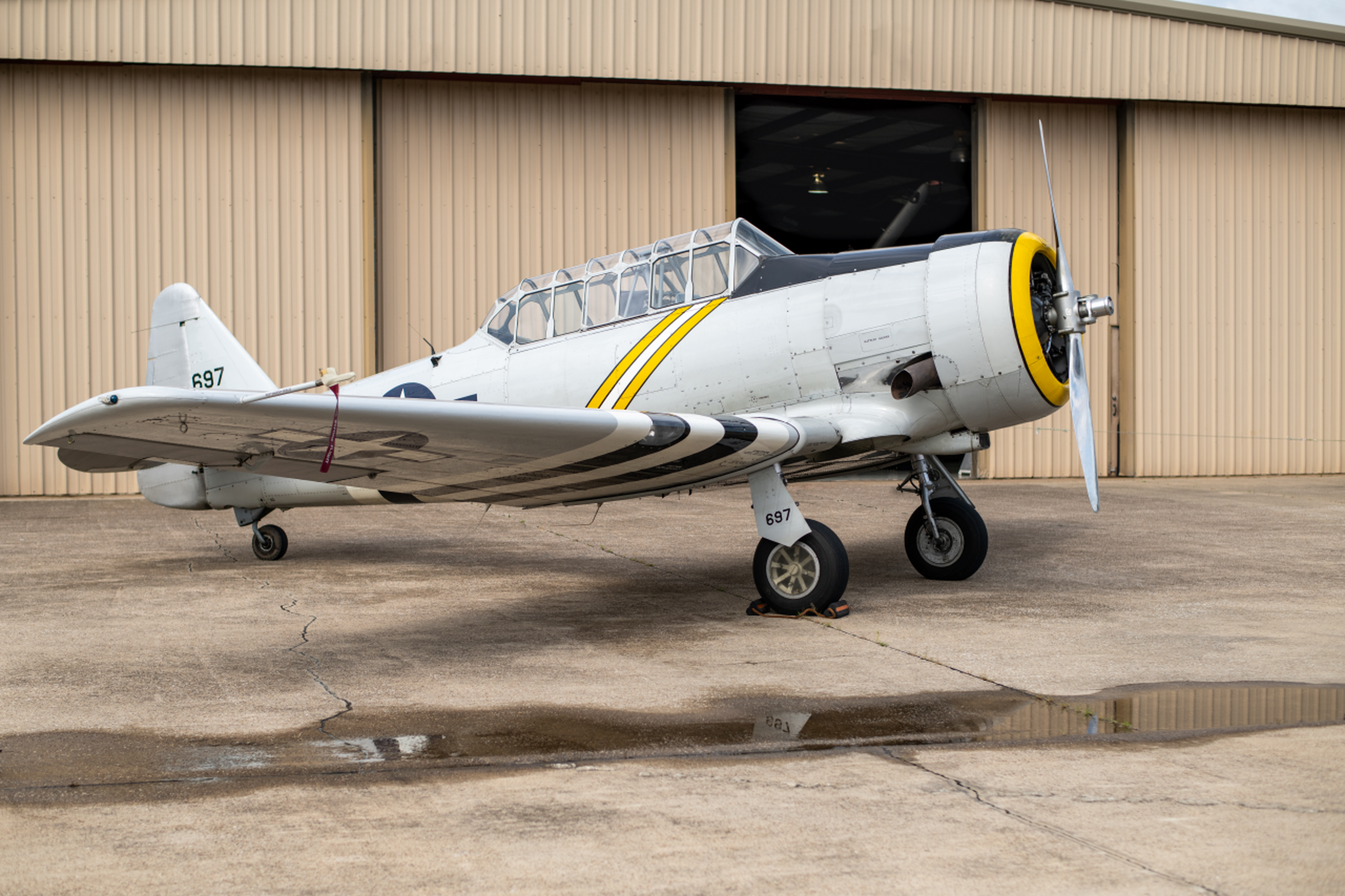 A vintage propeller plane with yellow and black stripes is parked on a tarmac near a beige hangar.