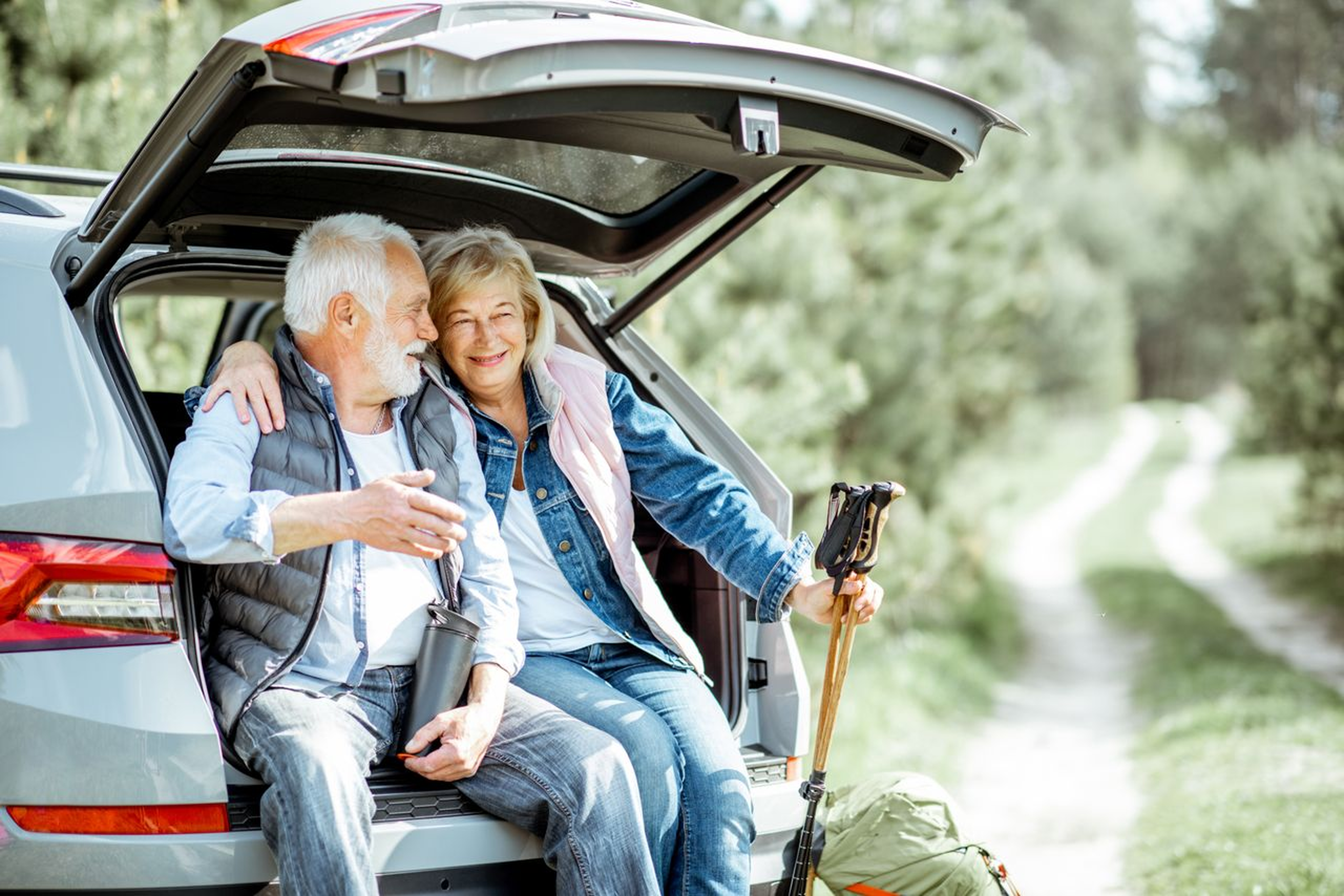 Elderly couple sitting in an SUV's open trunk, smiling and embracing, with a scenic forest path in the background.