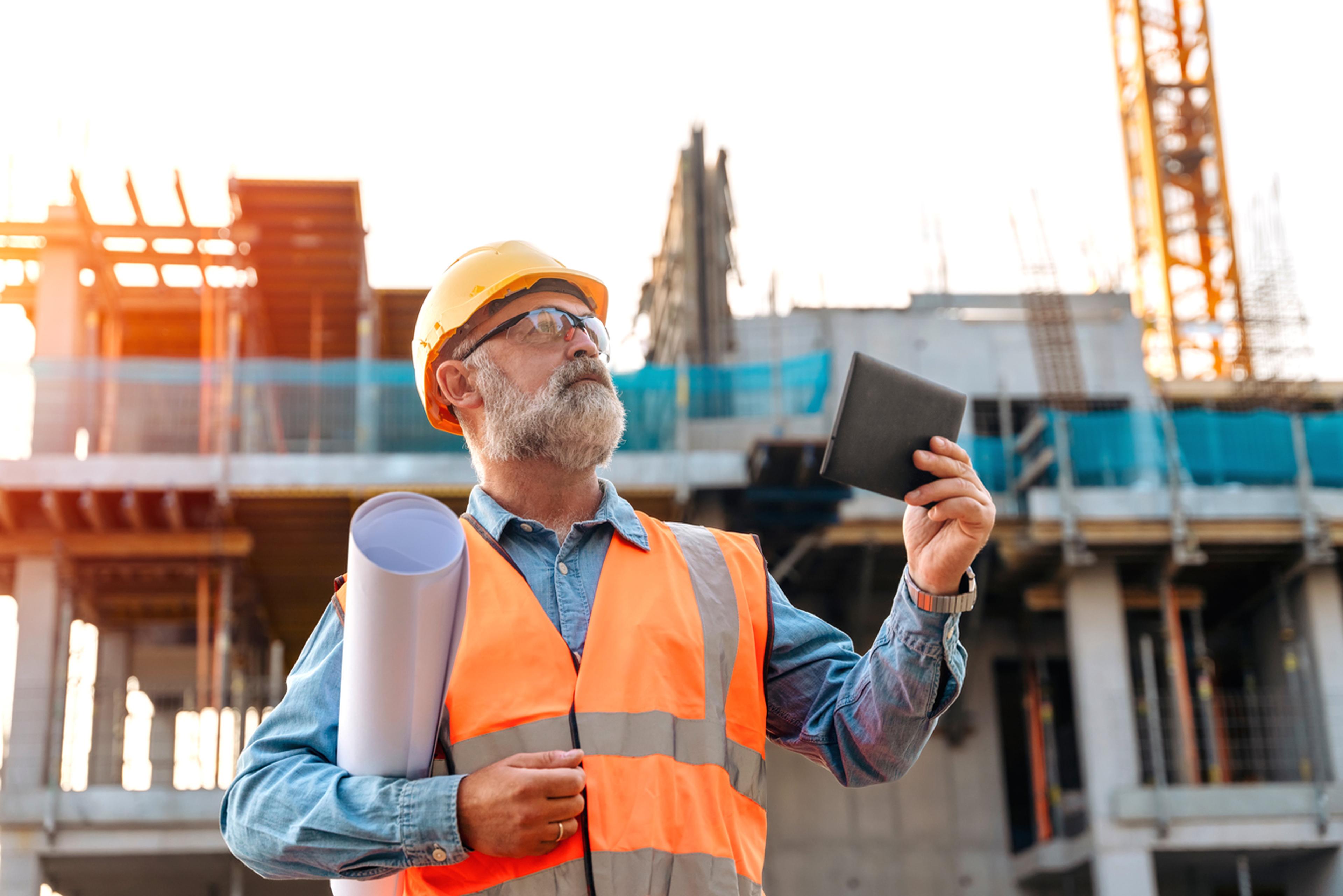 Bearded construction worker in hard hat and vest, holding blueprints and a tablet, inspects a building site with partially completed structure.