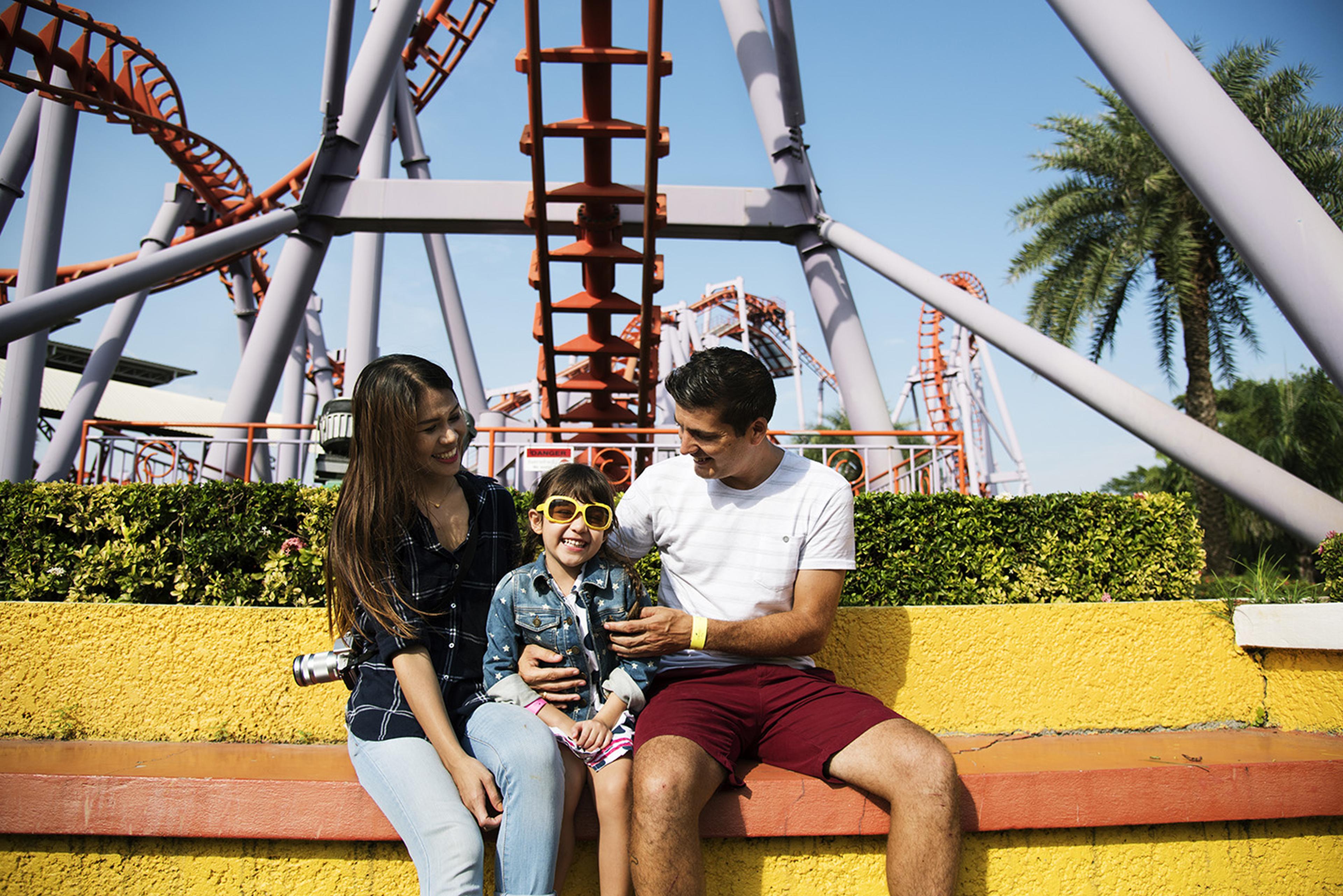 A family sits on a bench at an amusement park, with roller coasters in the background. The child wears yellow sunglasses and smiles happily.
