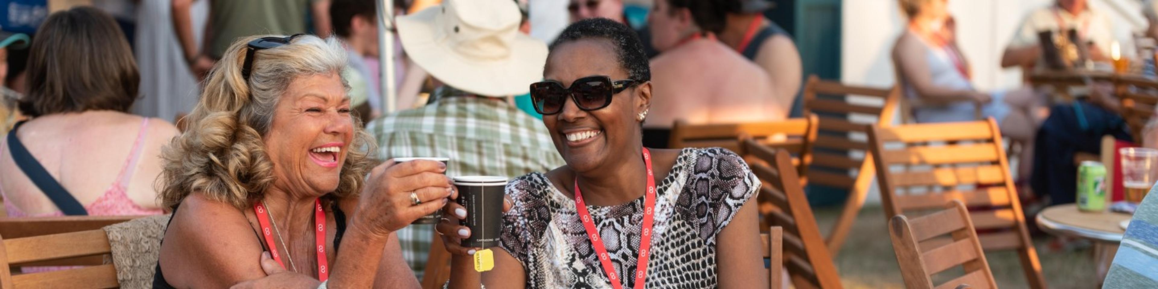 Two women sitting outdoors at a festival, laughing and holding drinks. People are seated on wooden chairs in the background.