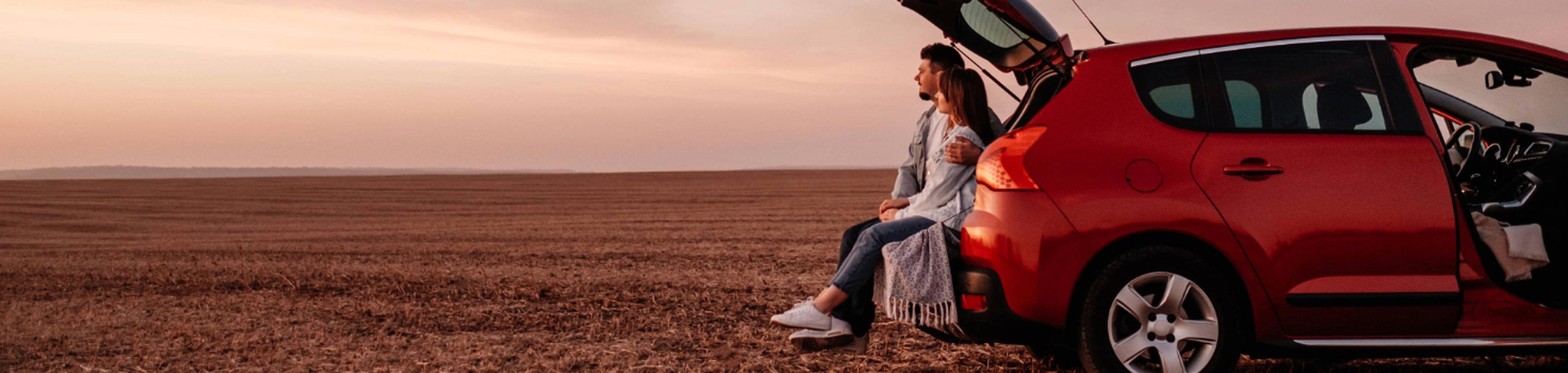 A couple sits in the open boot of a red car, gazing at a field during sunset