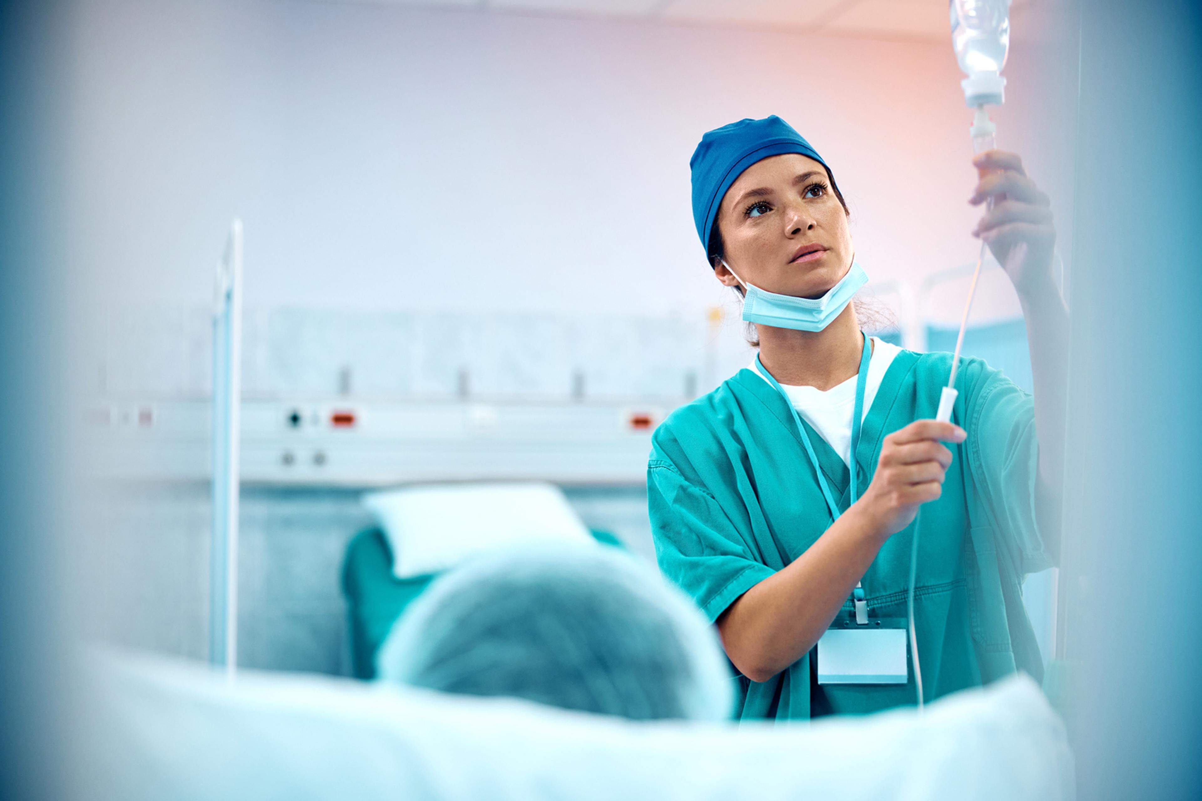 Nurse in scrubs adjusting an IV drip in a hospital room, with a masked face and focused expression.
