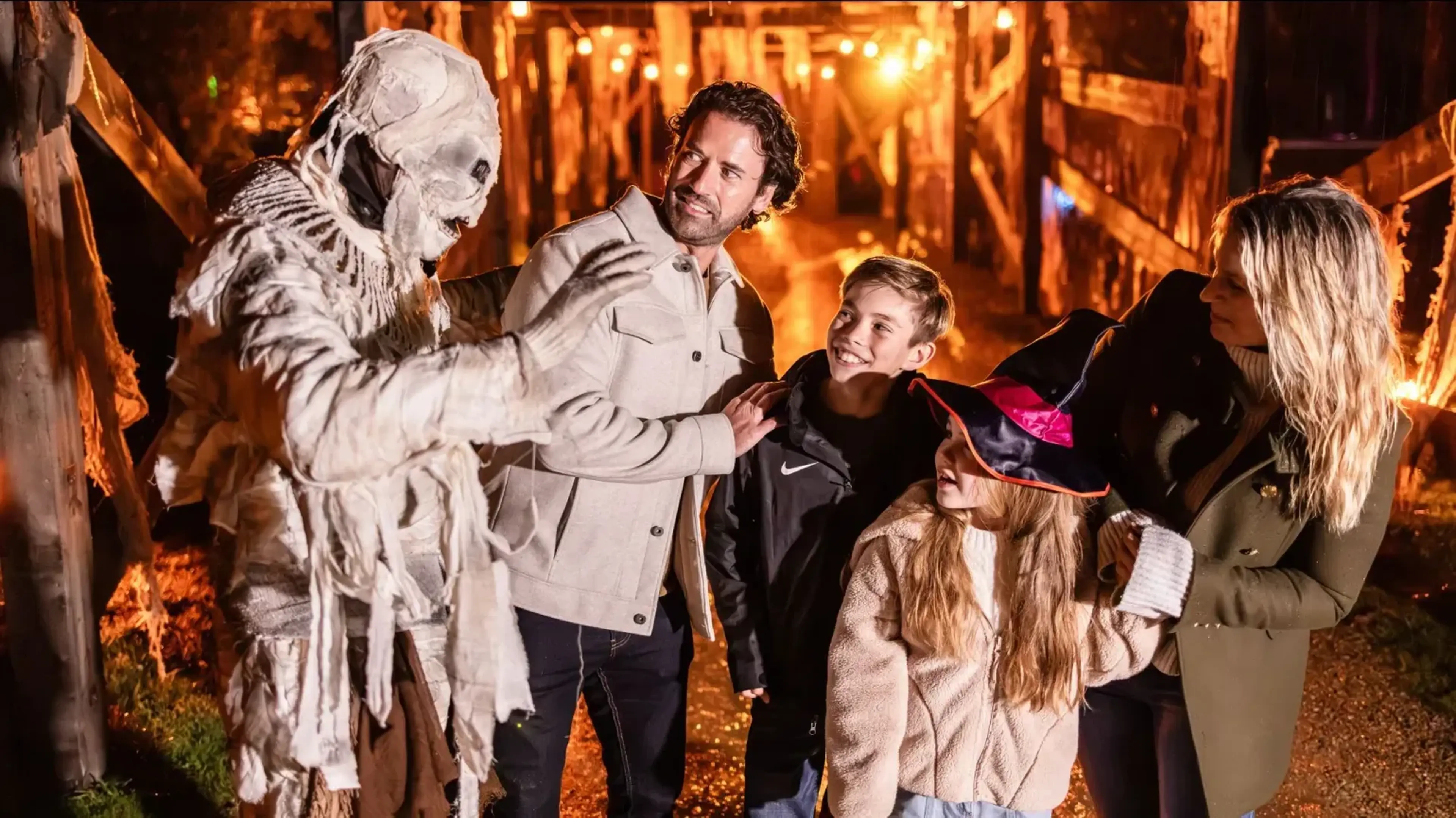 A family enjoys a spooky Halloween trail at night, encountering a mummy performer among glowing autumn lights and decorations.