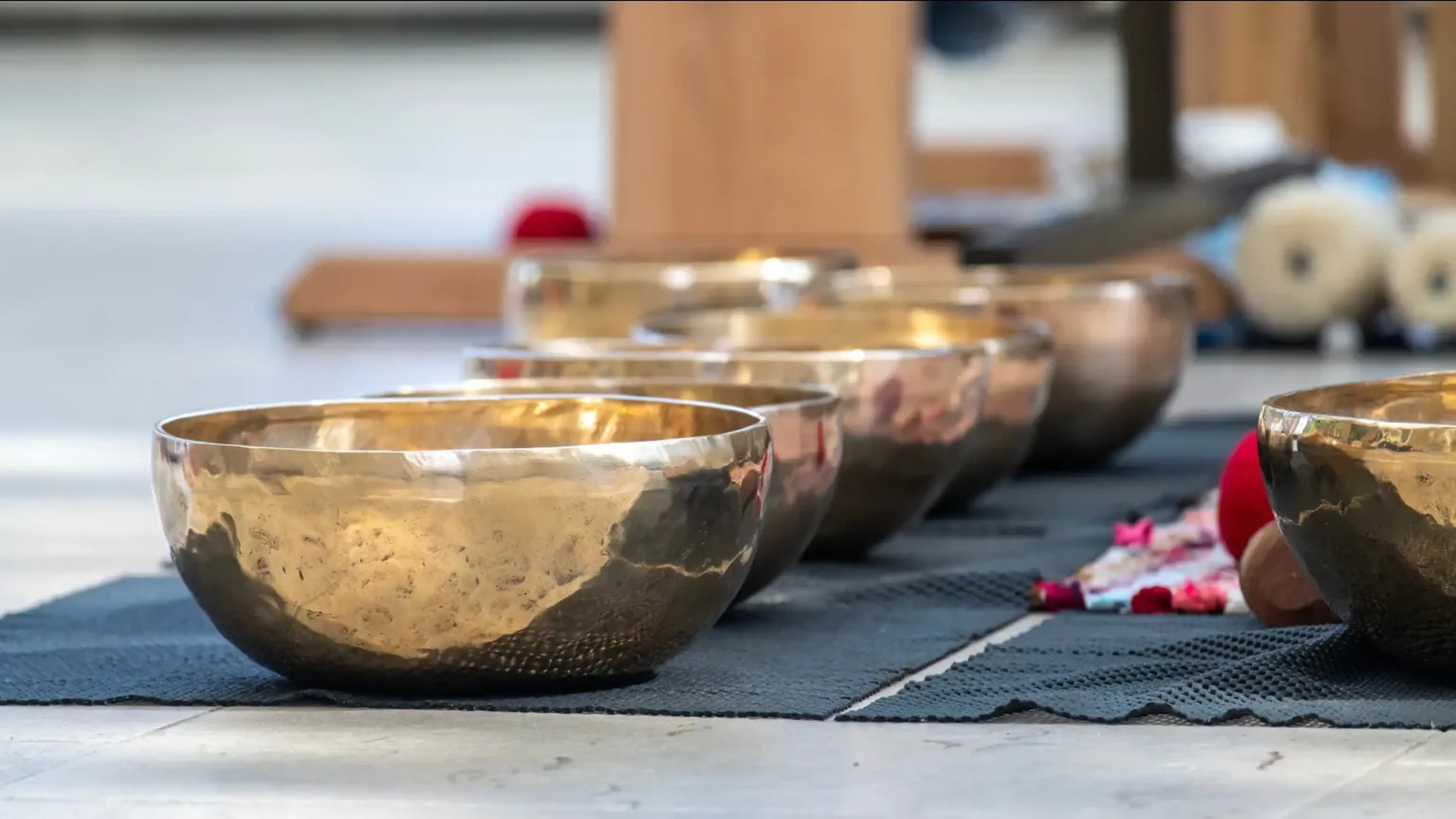 A row of shiny singing bowls arranged on a dark mat, with blurred background elements suggesting a serene setting.