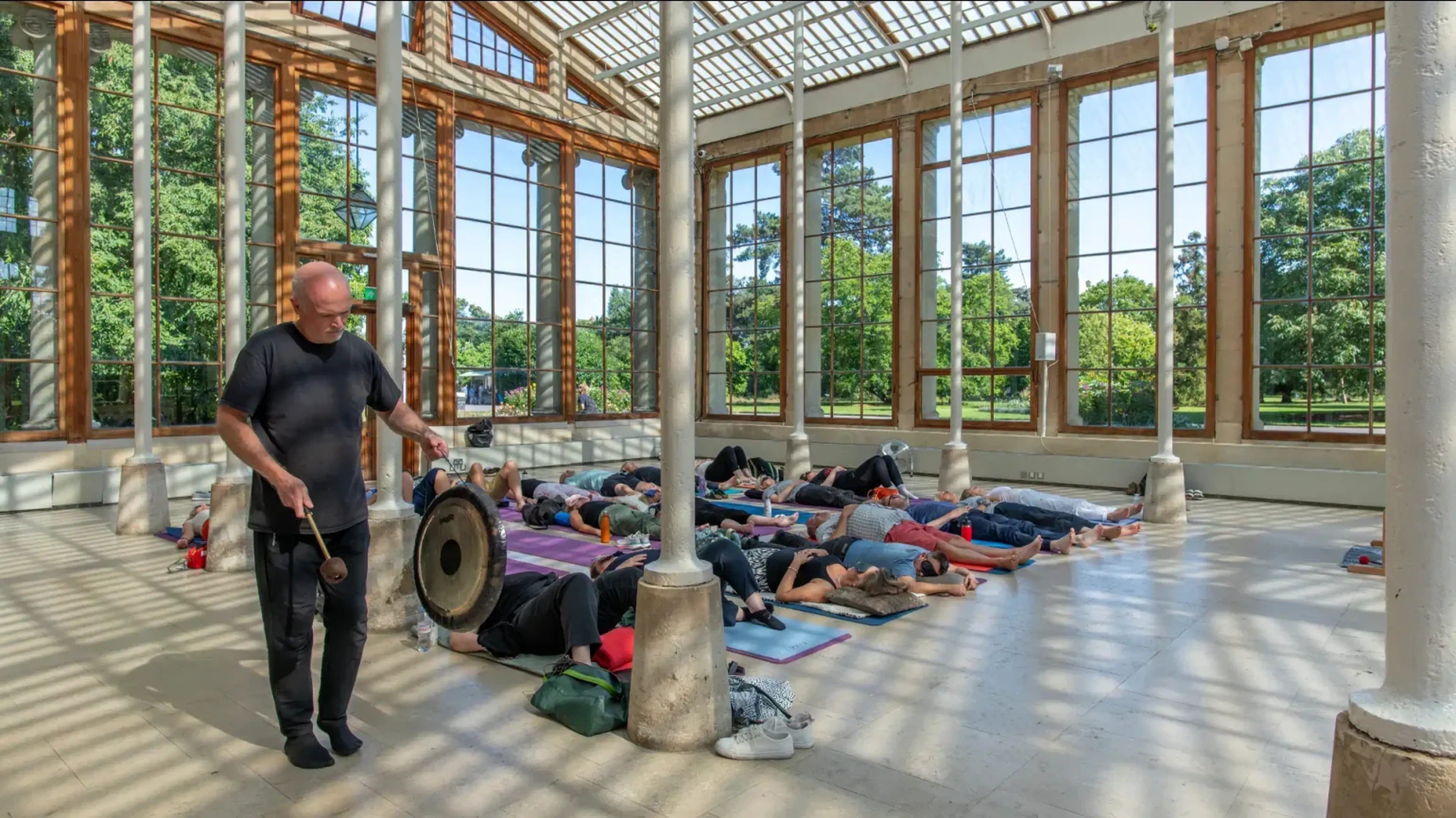 A person plays a gong in a sunlit glass pavilion while a group lies on yoga mats, relaxing.