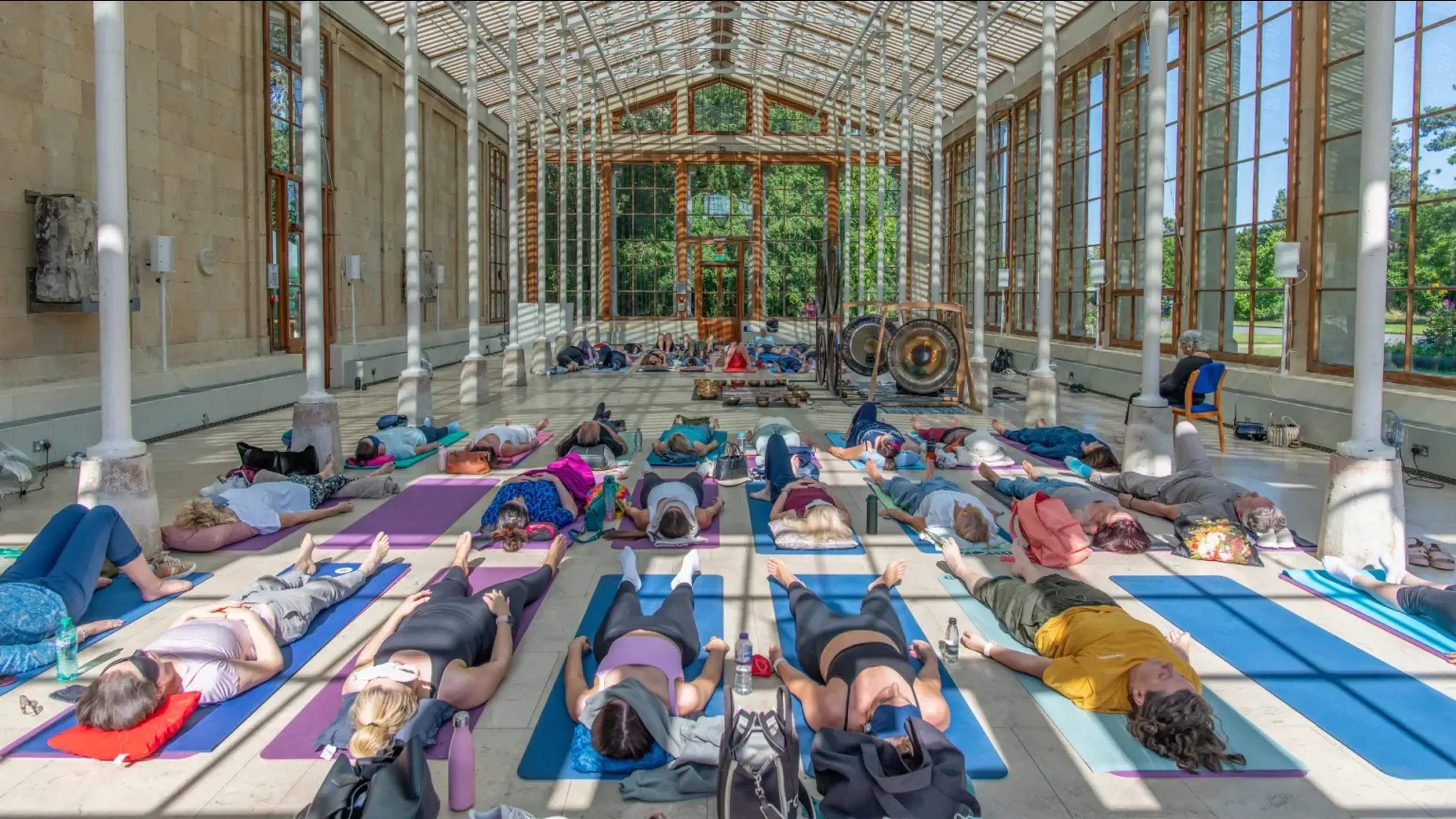 People lying on yoga mats in a bright hall with large windows, participating in a sound meditation session with gongs and instruments at the front.