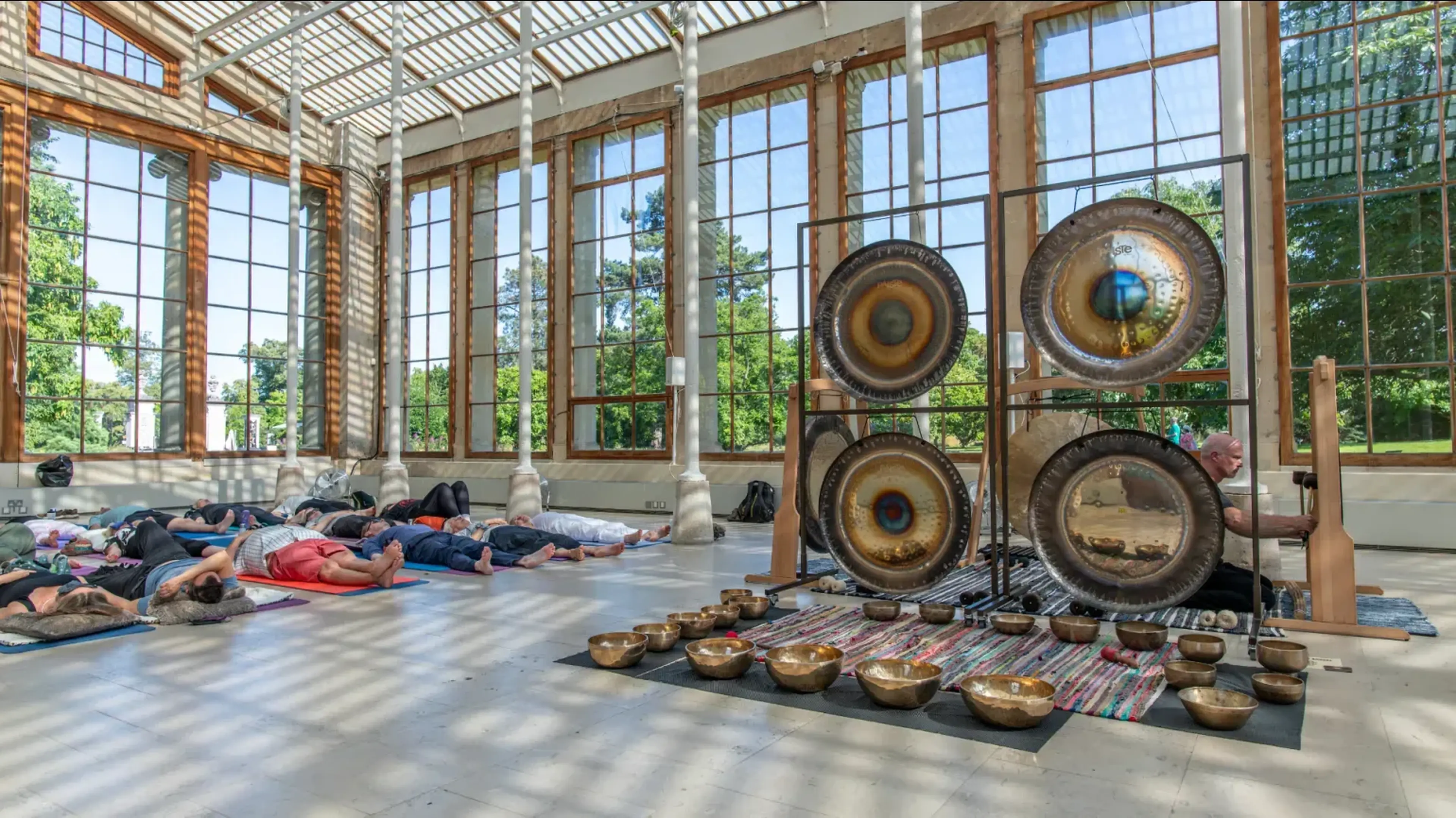 People lying on mats in a sunlit room with large windows, listening to a musician playing gongs and bowls for a sound meditation session.