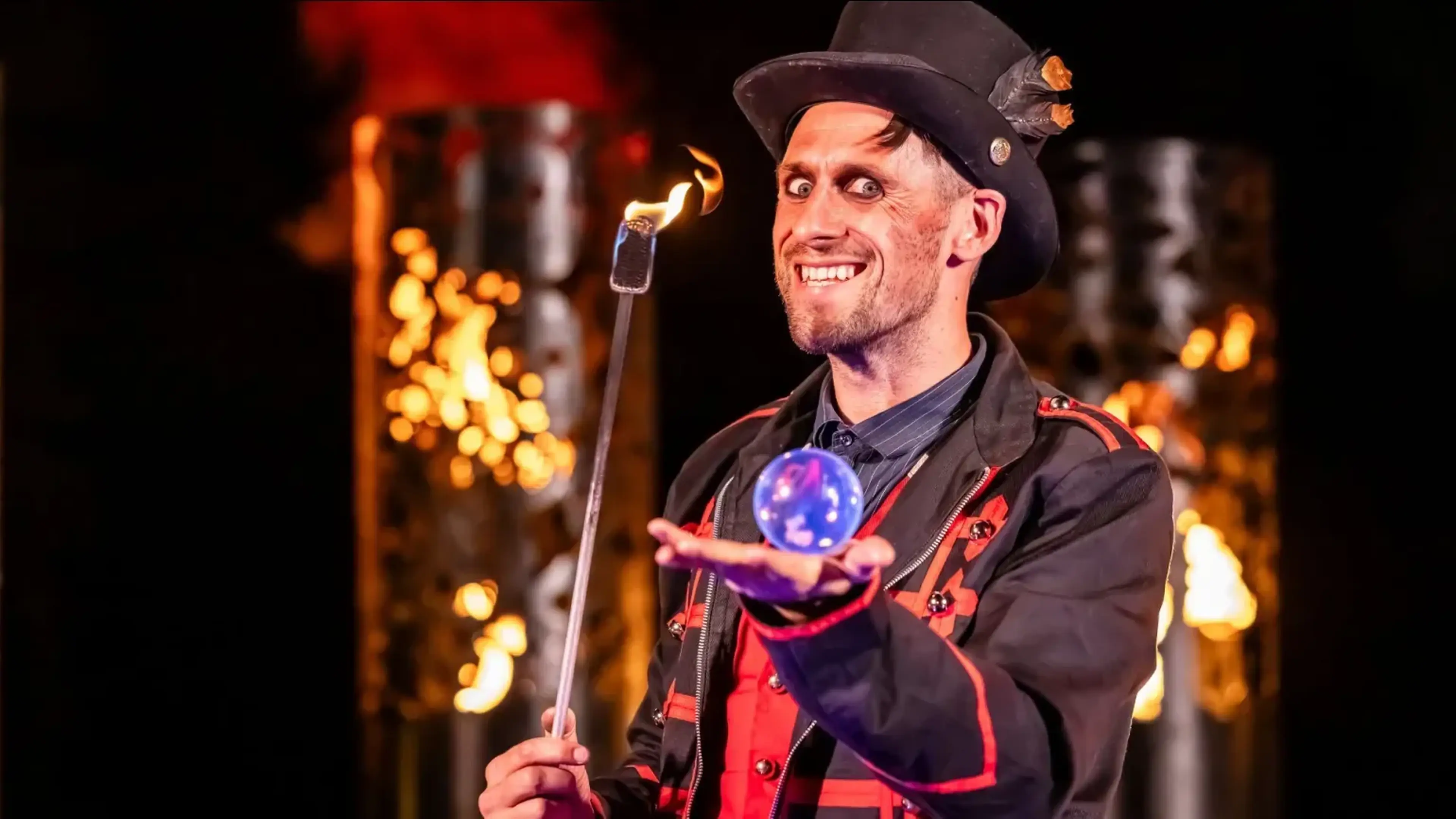 A smiling magician in a black-and-red costume and top hat holds a glowing blue crystal ball and a flaming wand during a Halloween-themed performance.
