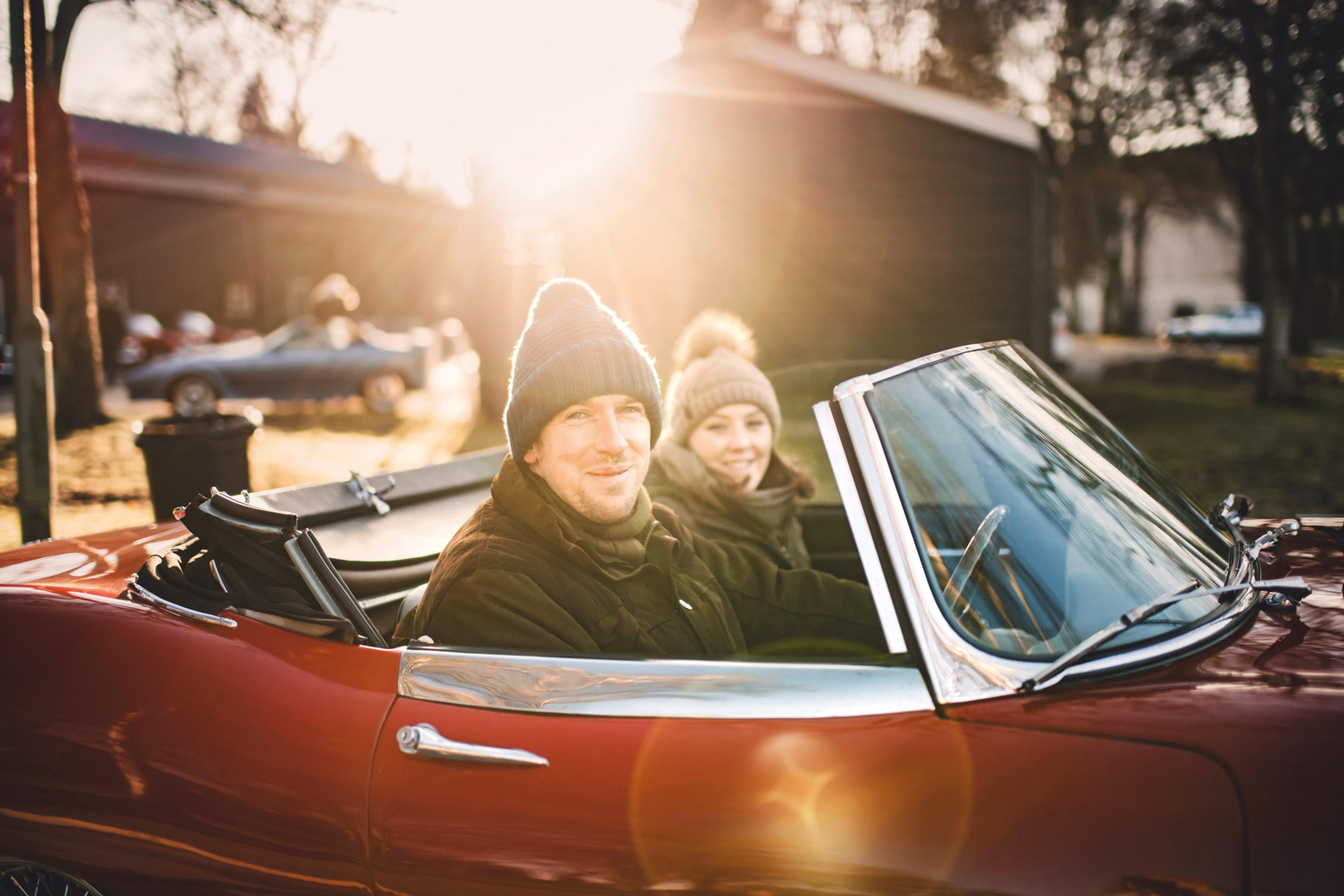 A couple in winter clothing enjoys a sunny drive in a red convertible. Sunlight shines brightly in the background.