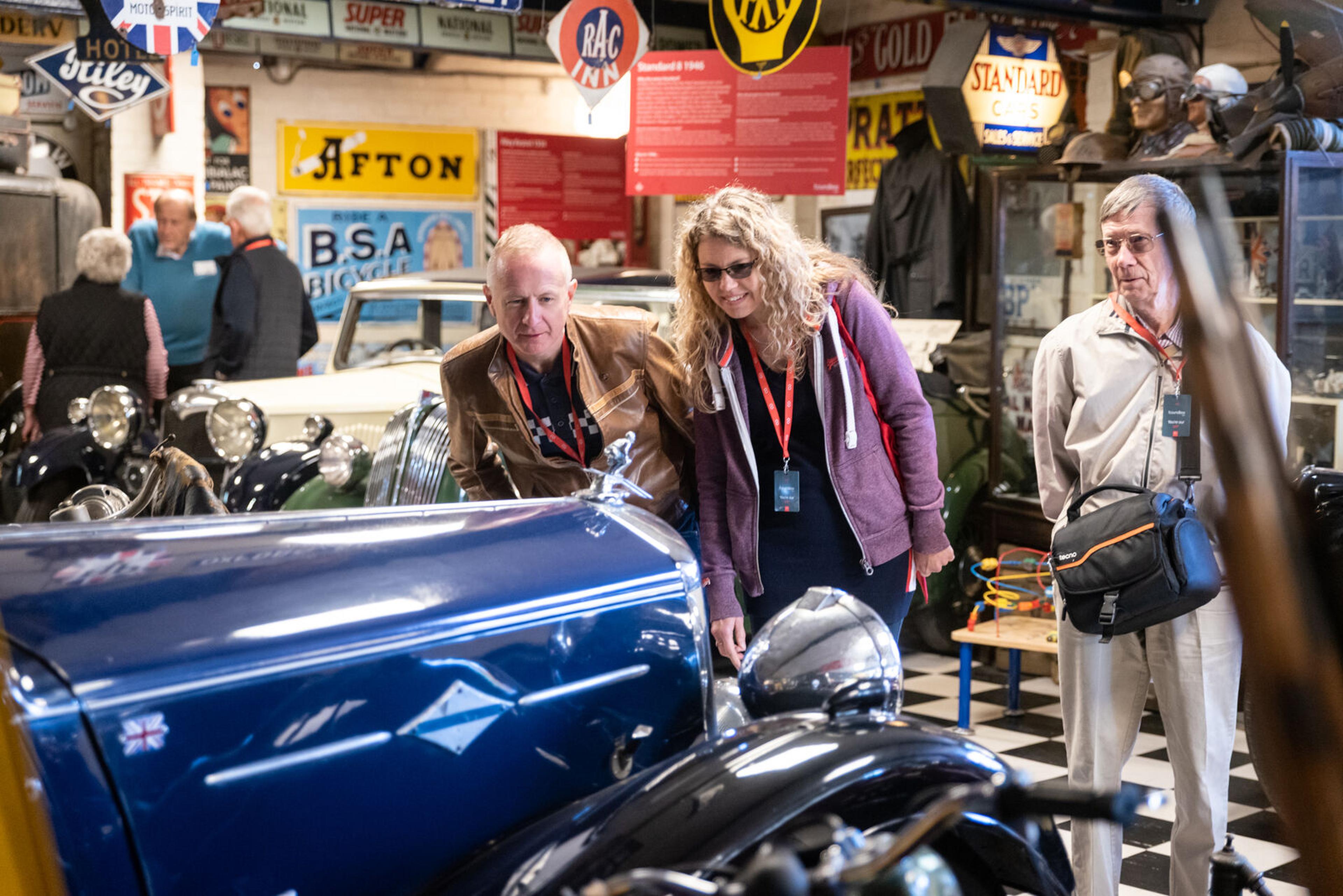Visitors admire a classic blue car in a vintage automobile museum, surrounded by retro signage and memorabilia.