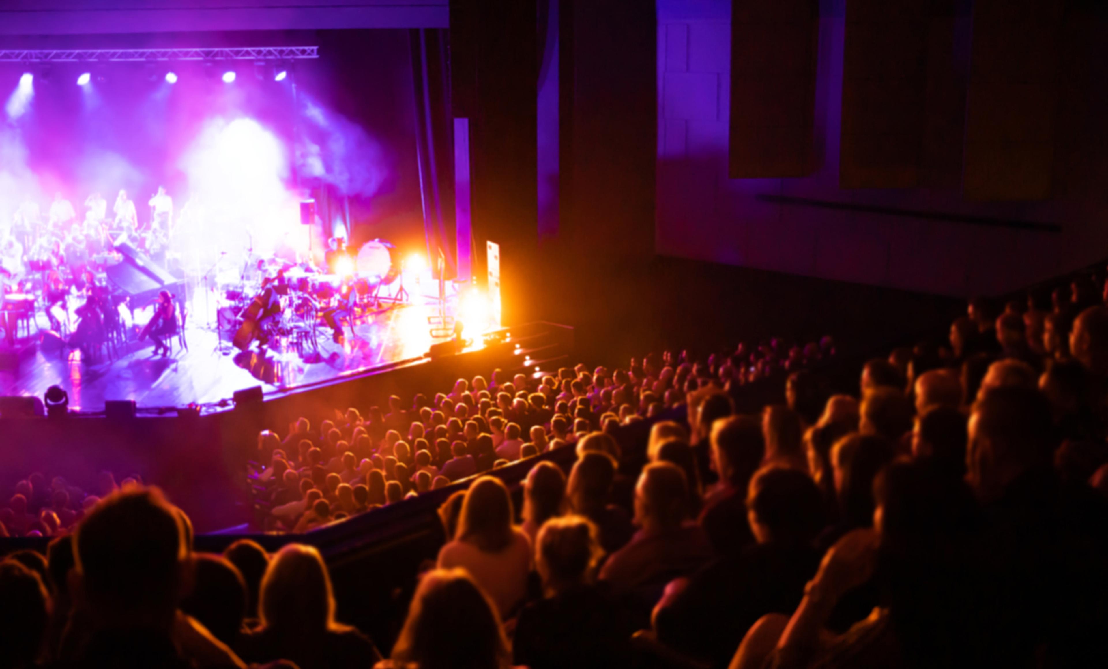 A packed theater with an audience watching a live stage performance, illuminated by vibrant purple and orange lights.