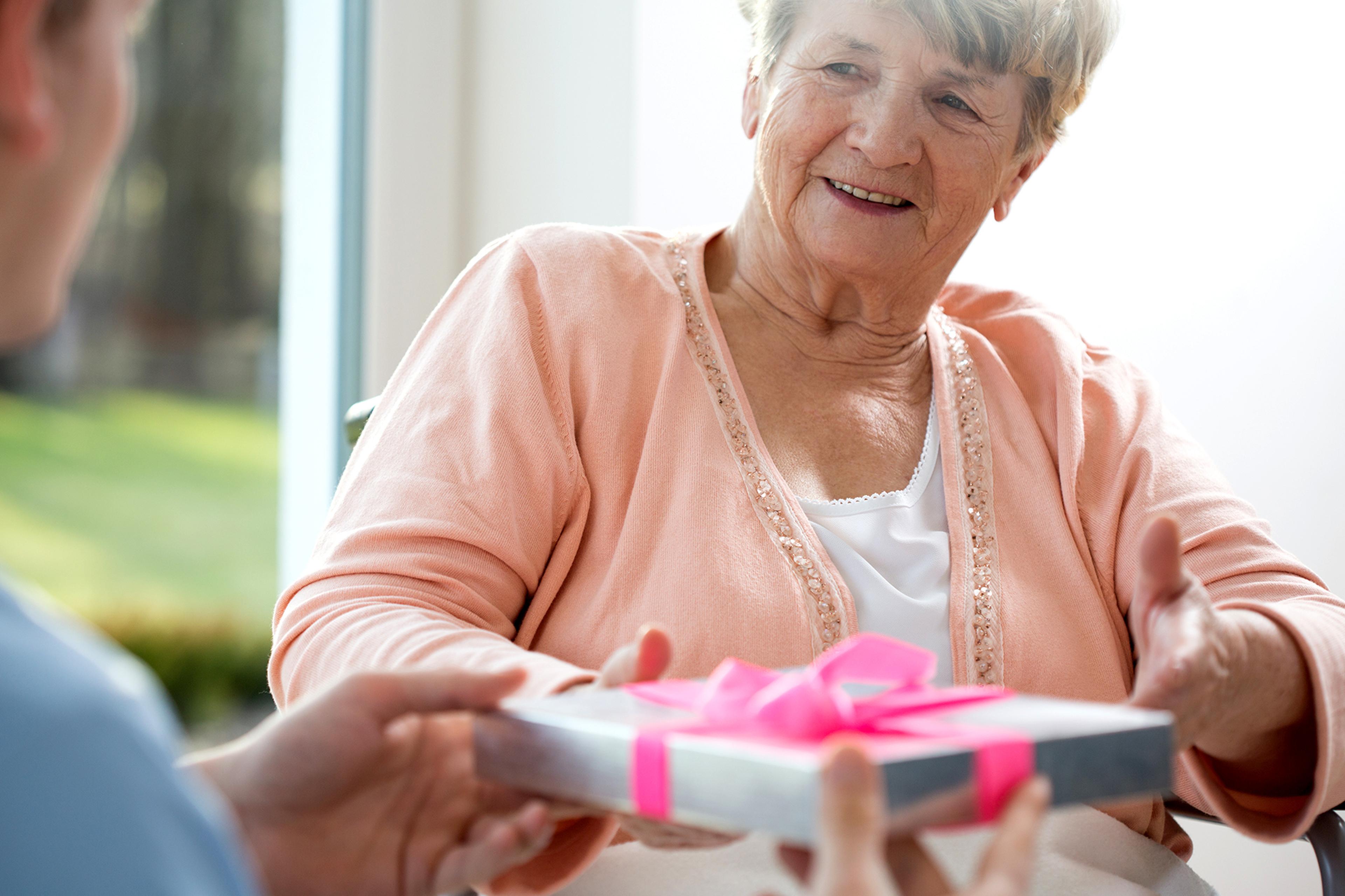 Elderly woman smiling while receiving a wrapped gift with a pink ribbon from another person indoors.