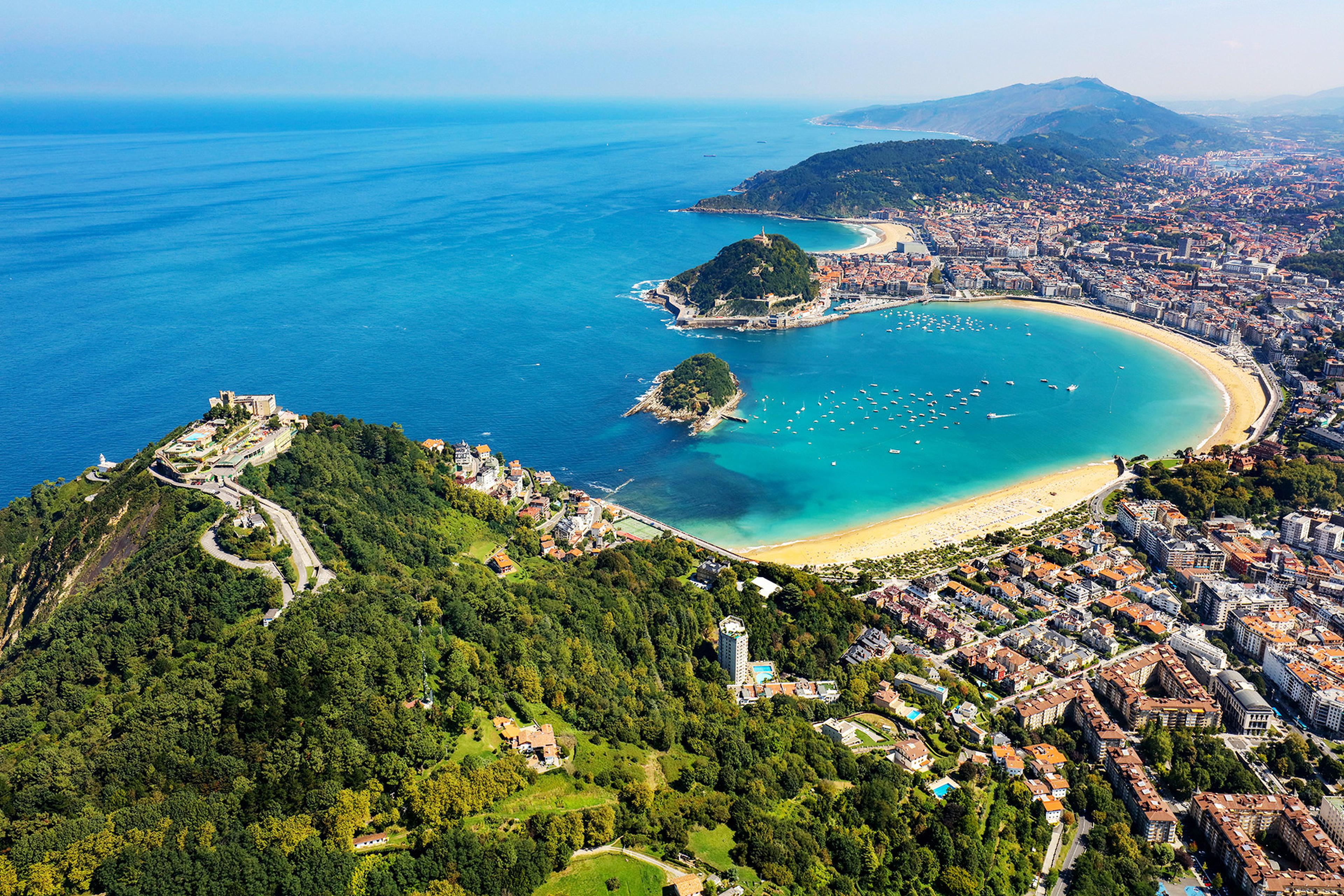 Aerial view of a coastal city with a crescent-shaped bay, sandy beaches, green hills, and boats dotting the turquoise water.