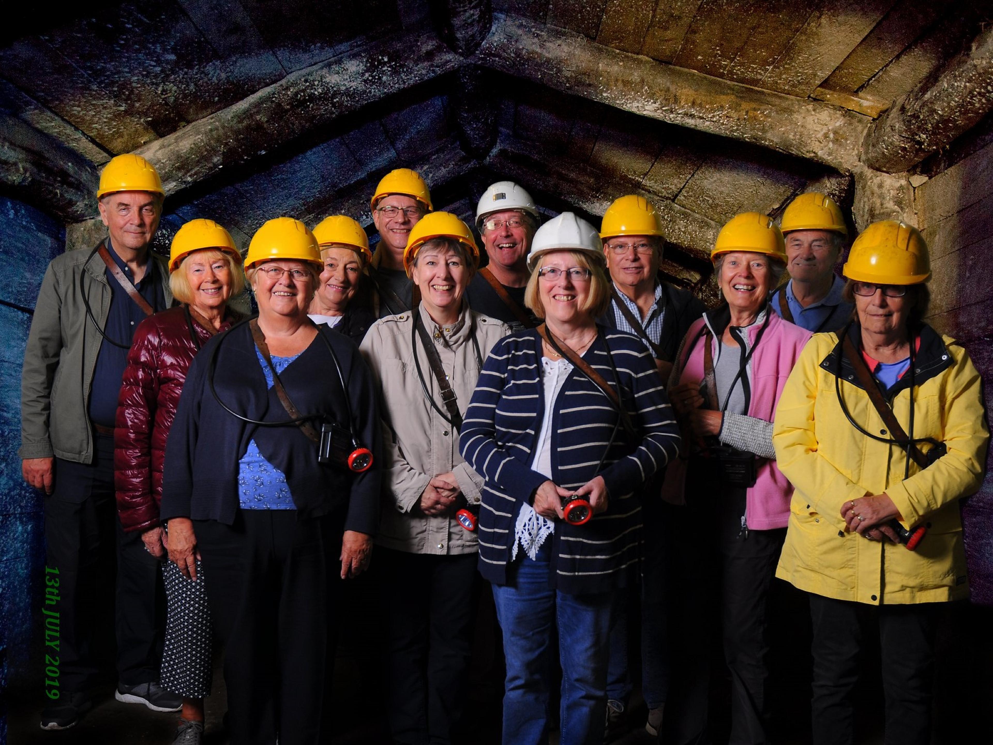 Group of people wearing hard hats and holding flashlights pose in a tunnel with wooden beams overhead.