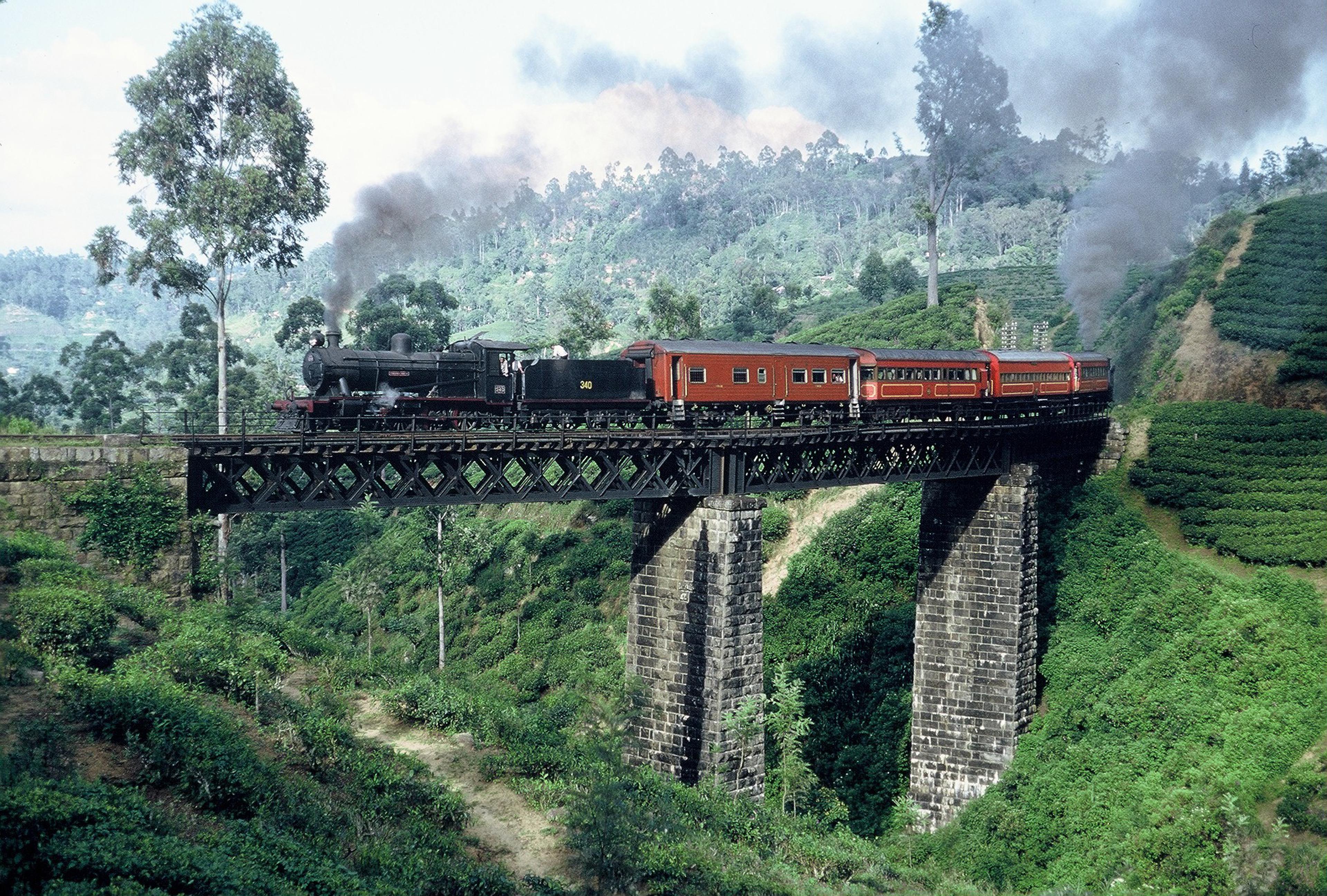 This photograph captures a steam train crossing the Nine Arches Bridge in Sri Lanka’s highlands, surrounded by lush tea plantations and misty hills. The bridge, officially known as the Demodara Nine Arch Bridge, is a colonial-era engineering marvel that carries the Ella–Demodara railway line across a deep ravine.