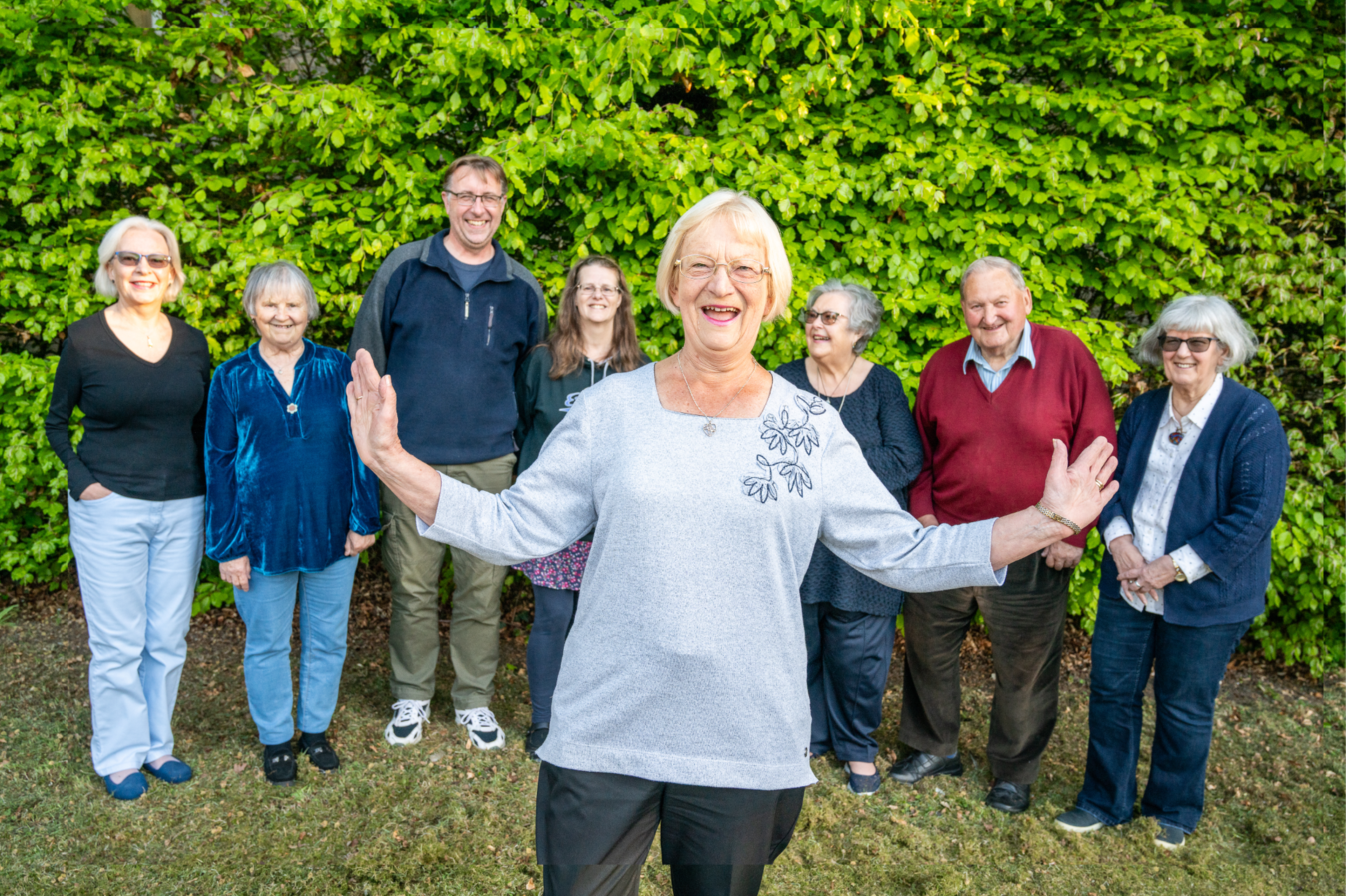A smiling woman in a gray sweater stands with her arms open in front of a happy group of people, with lush green foliage in the background.