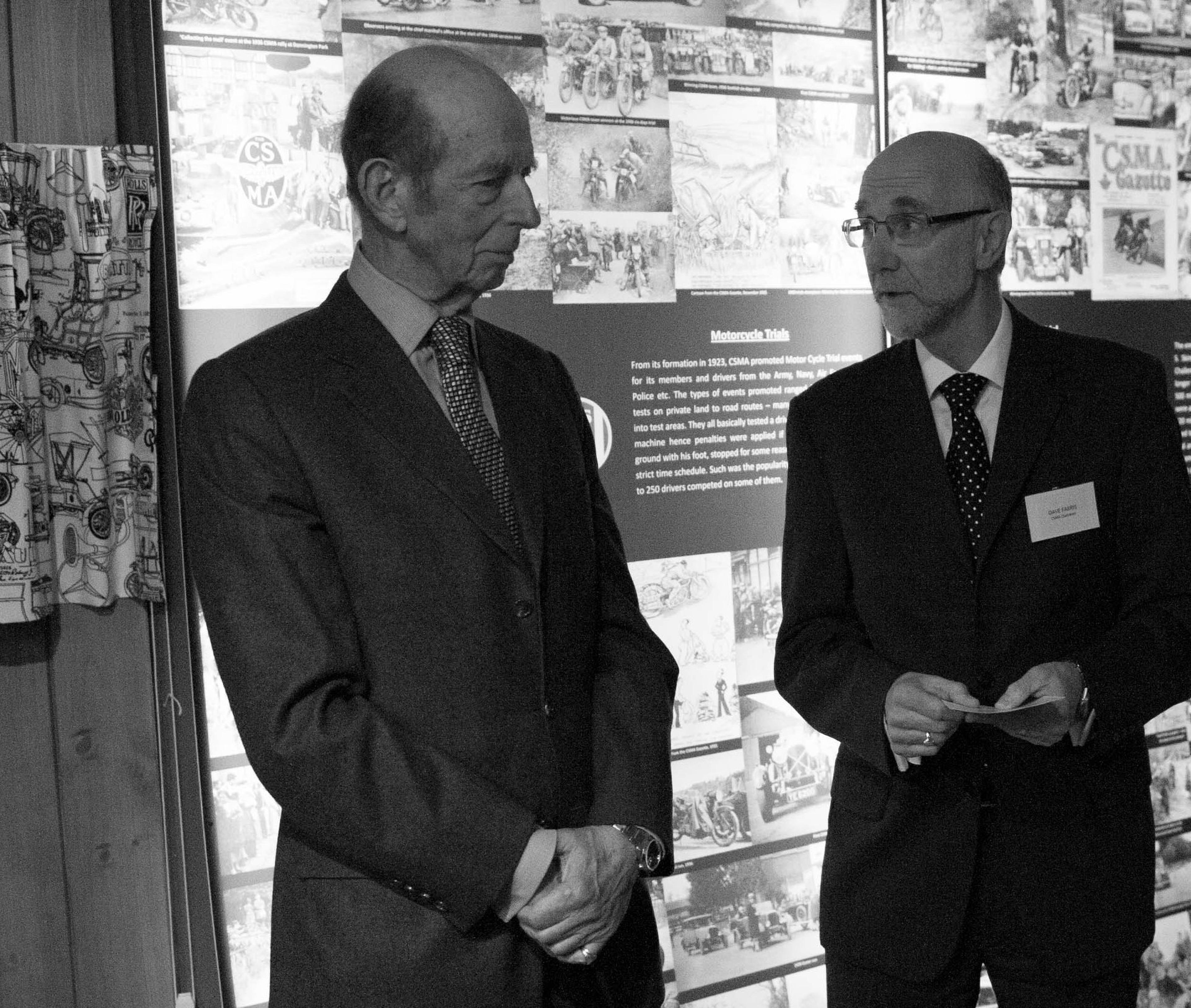 Black-and-white photo of two suited men conversing at a historical exhibition, surrounded by archival images and informational panels.