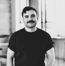 Black and white portrait of a smiling person with a mustache, wearing a dark T-shirt, standing in a softly lit room.