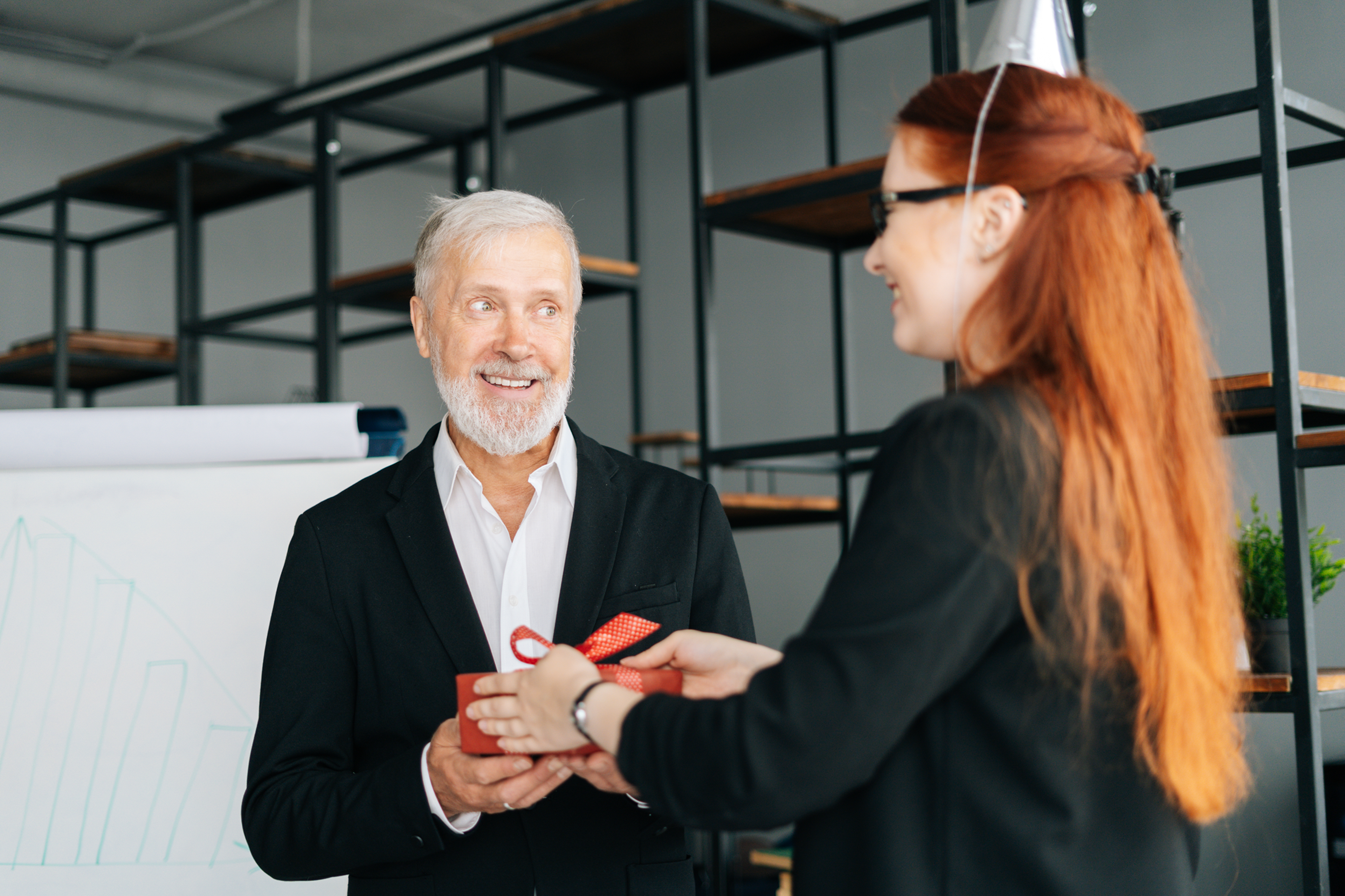 Woman gives a gift to a smiling older man in an office setting, with shelves and a whiteboard in the background.