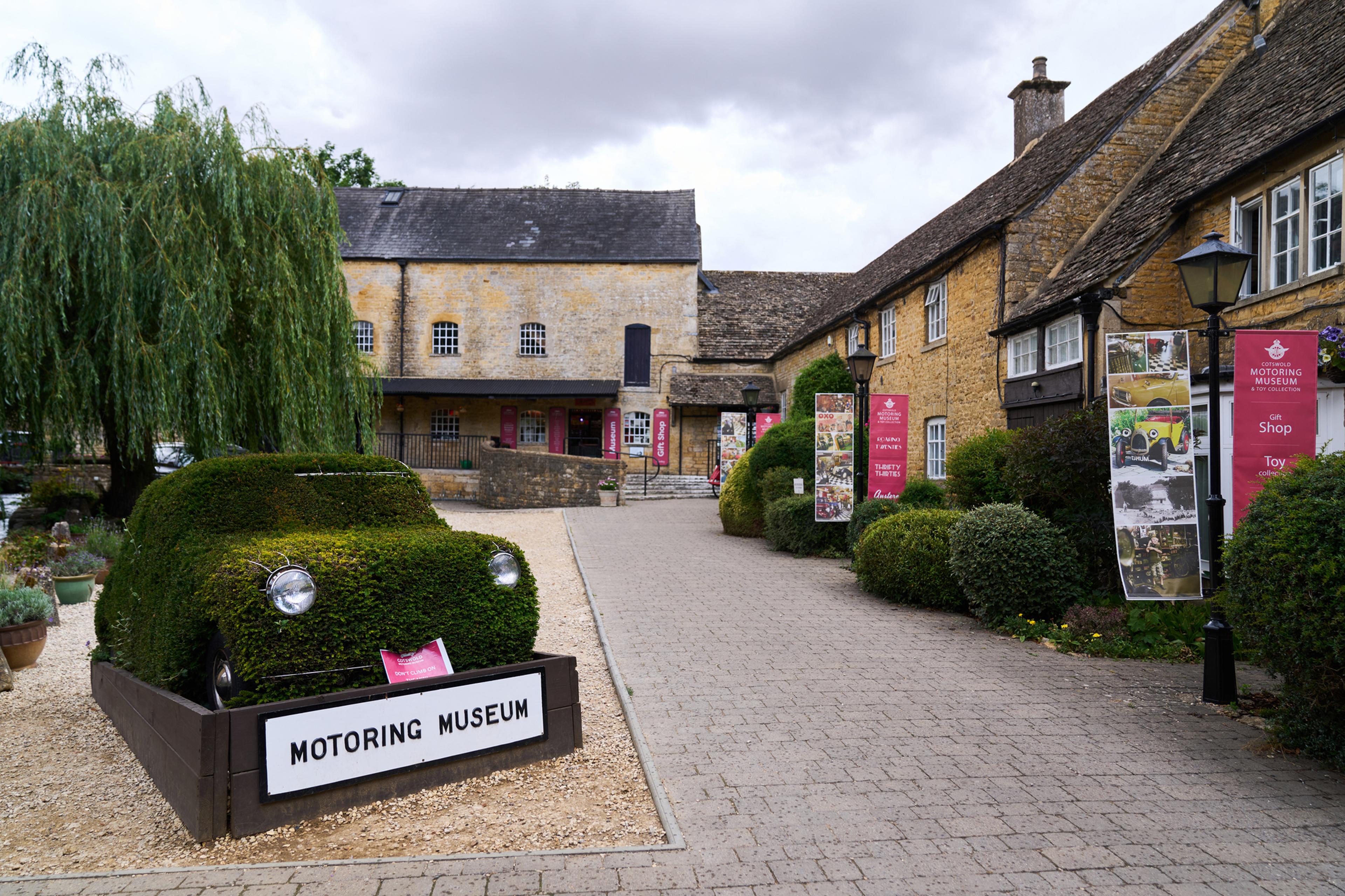 Entrance to a motoring museum with a car-shaped bush, cobblestone path, and historic stone buildings, under a cloudy sky.